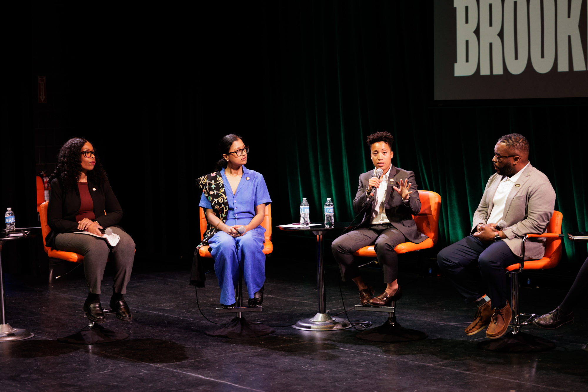 Four people sit on stage in orange chairs, engaged in a panel discussion. One person speaks into a microphone while the others listen. A "BROOK" sign is partially visible in the background.