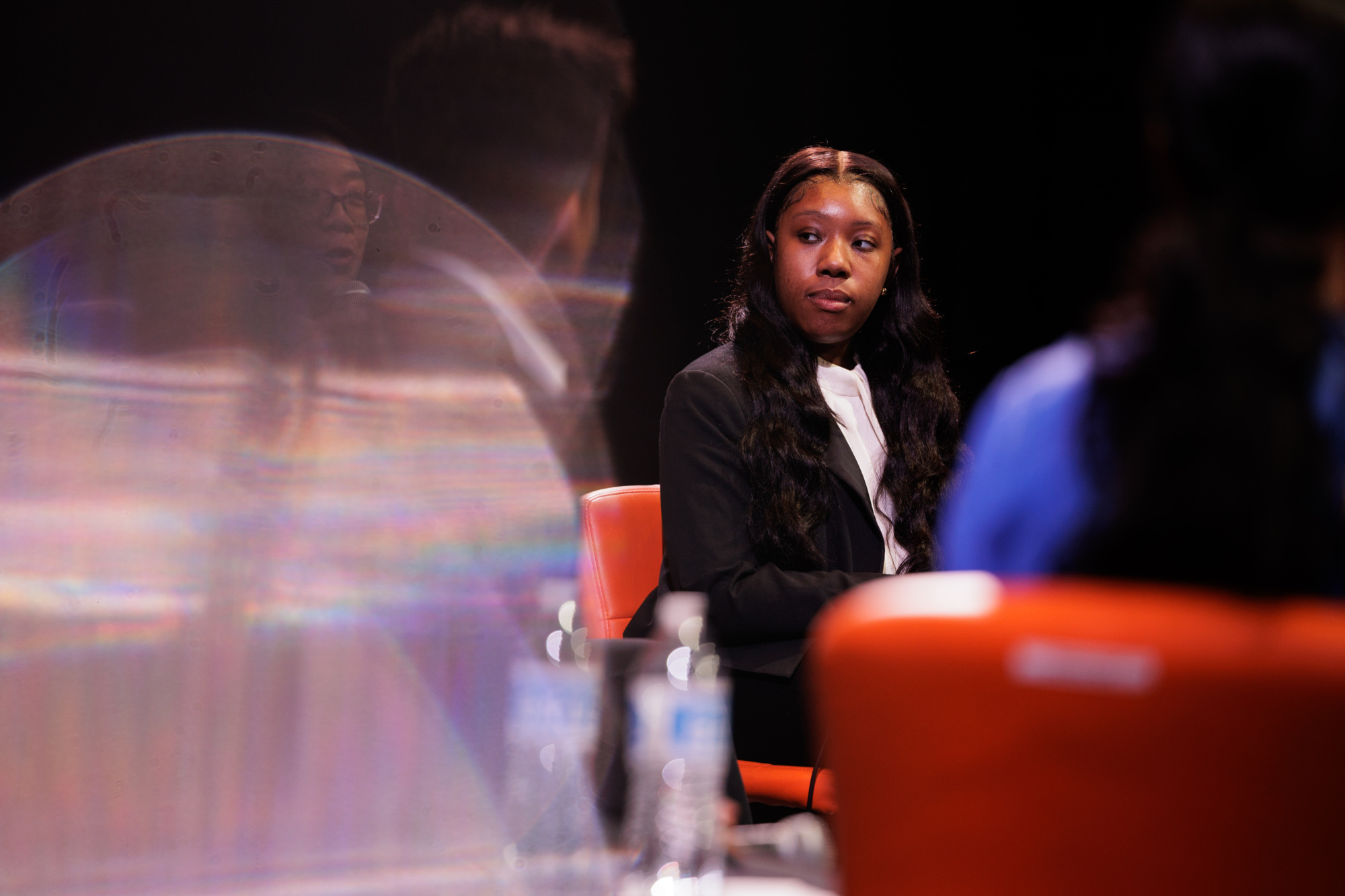 A woman in a black blazer sits on an orange chair during a panel discussion, with two other people and water bottles visible in the foreground.