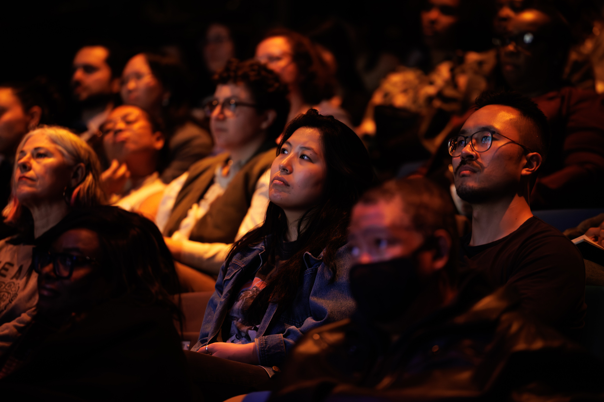 A diverse group of people sit closely together in a dimly lit auditorium, attentively watching something off-camera.