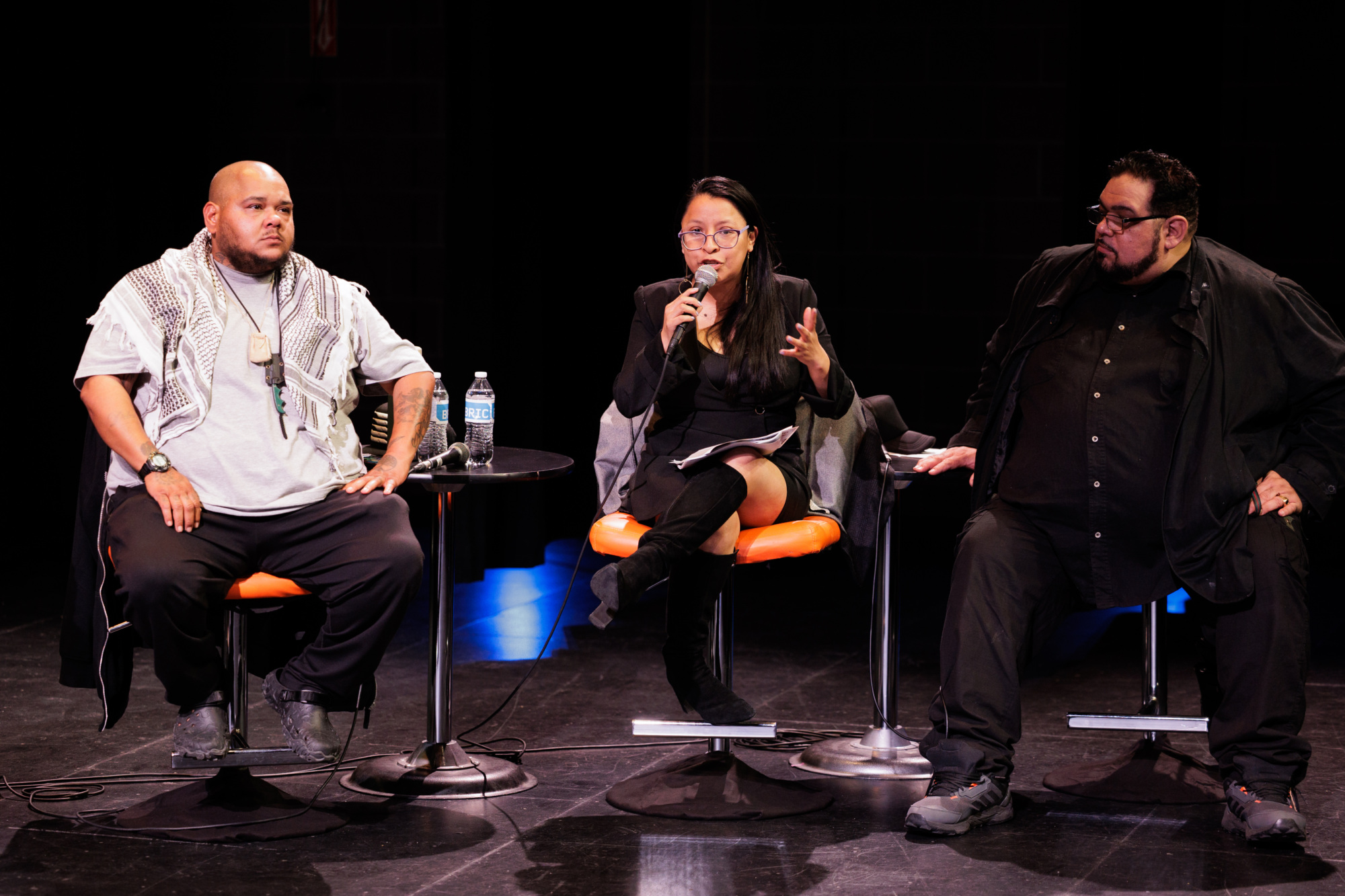 Three people sit on stage during a panel discussion; the woman in the center speaks into a microphone while the two men listen.