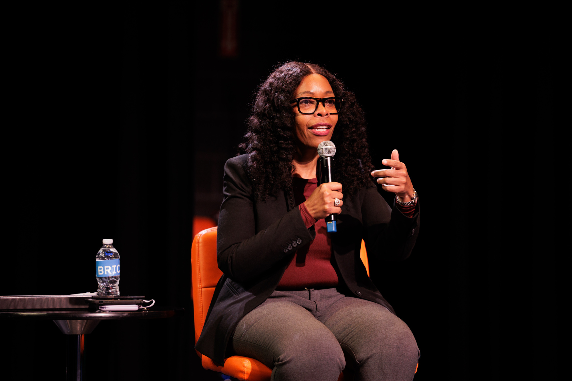 A woman with long curly hair and glasses speaks into a microphone while seated on an orange chair, with a water bottle on a small table beside her.