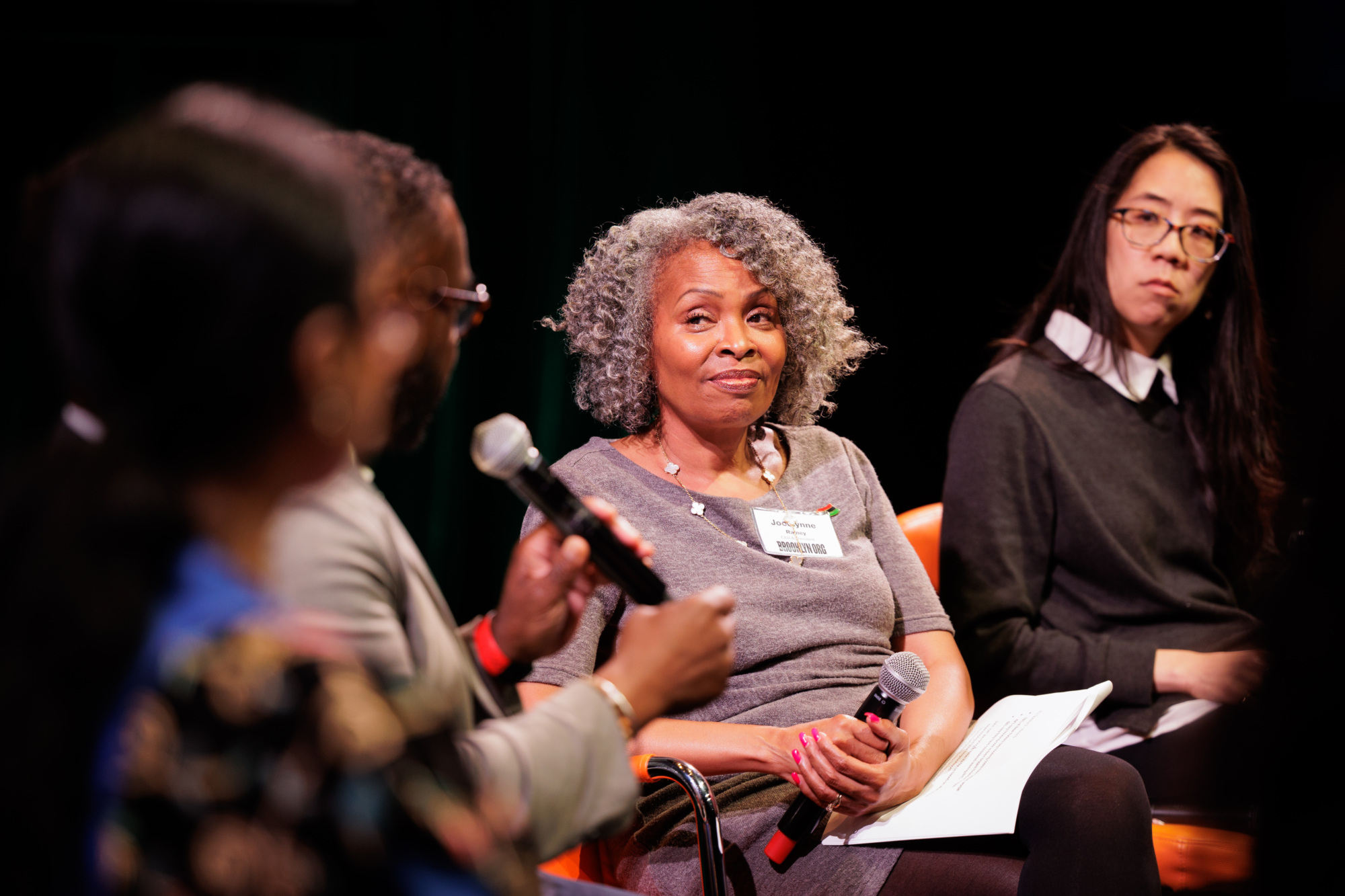 Four people sit on stage in a panel discussion; one woman is speaking into a microphone while the others listen attentively.