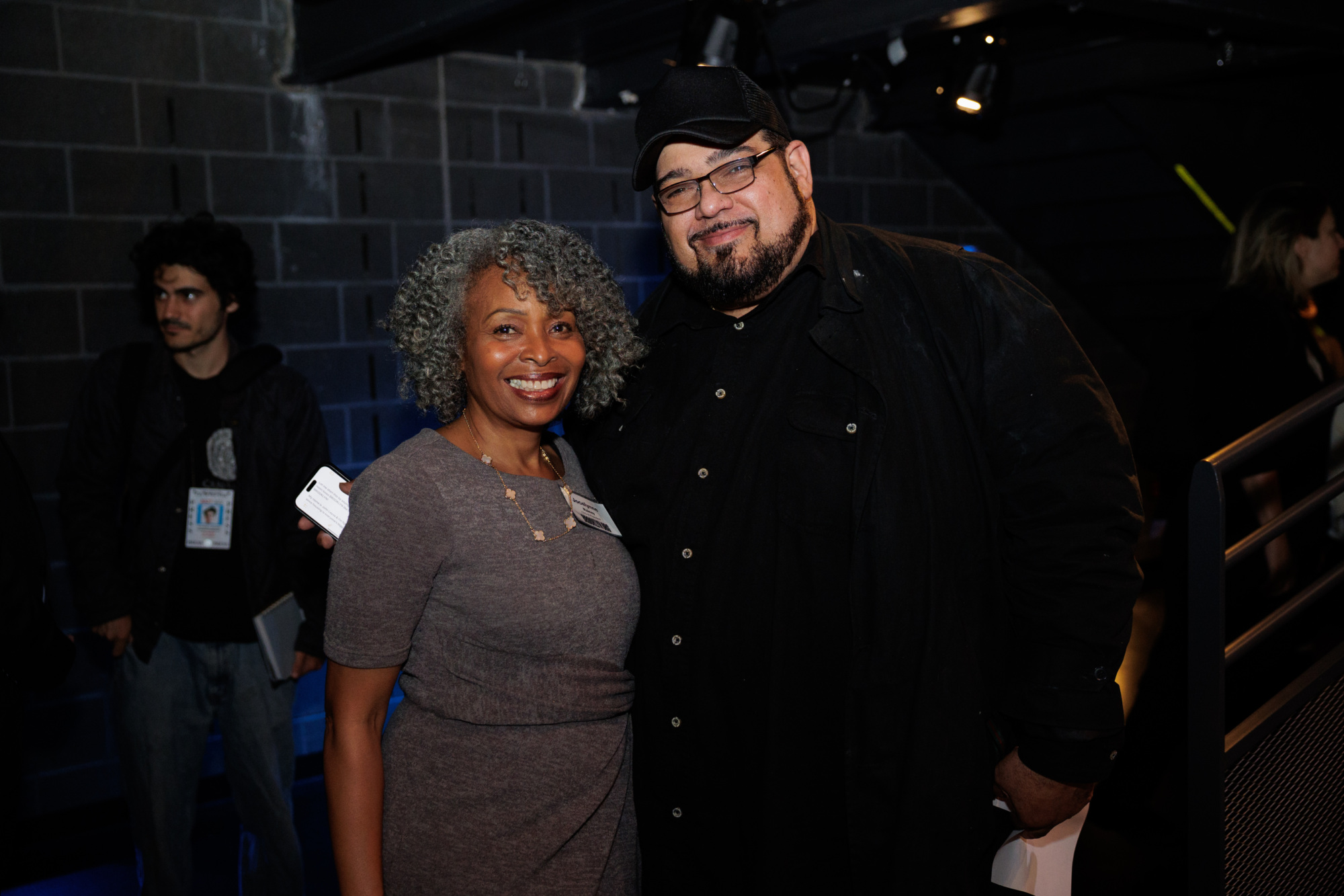 Two people stand side by side smiling at the camera at an indoor event, with other attendees visible in the background.