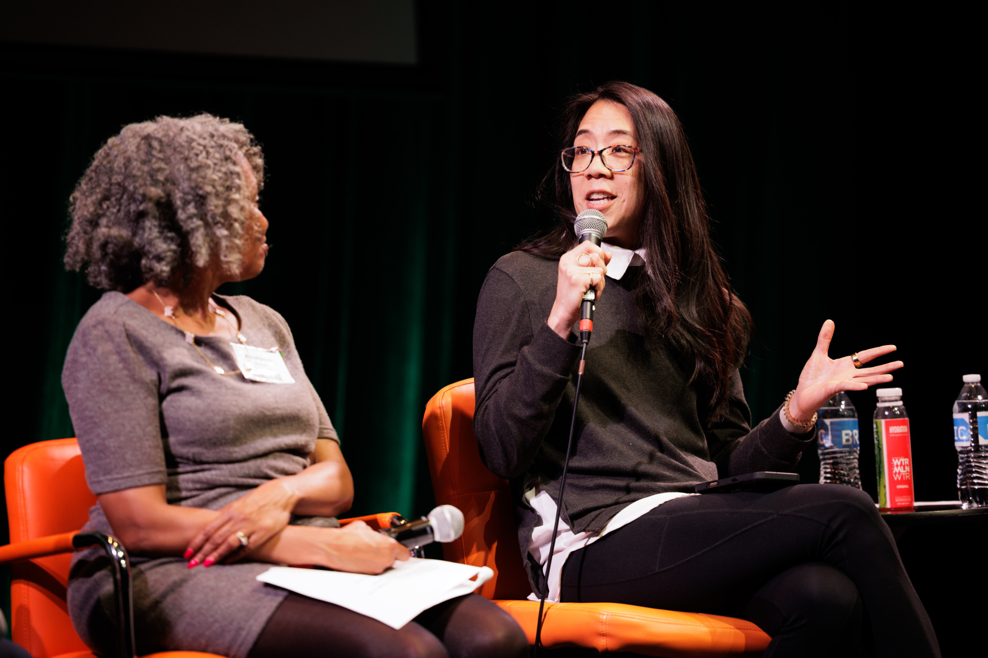 Two women sit on orange chairs during a panel discussion; one speaks into a microphone while the other listens. Bottled drinks and notepads are on the table between them.