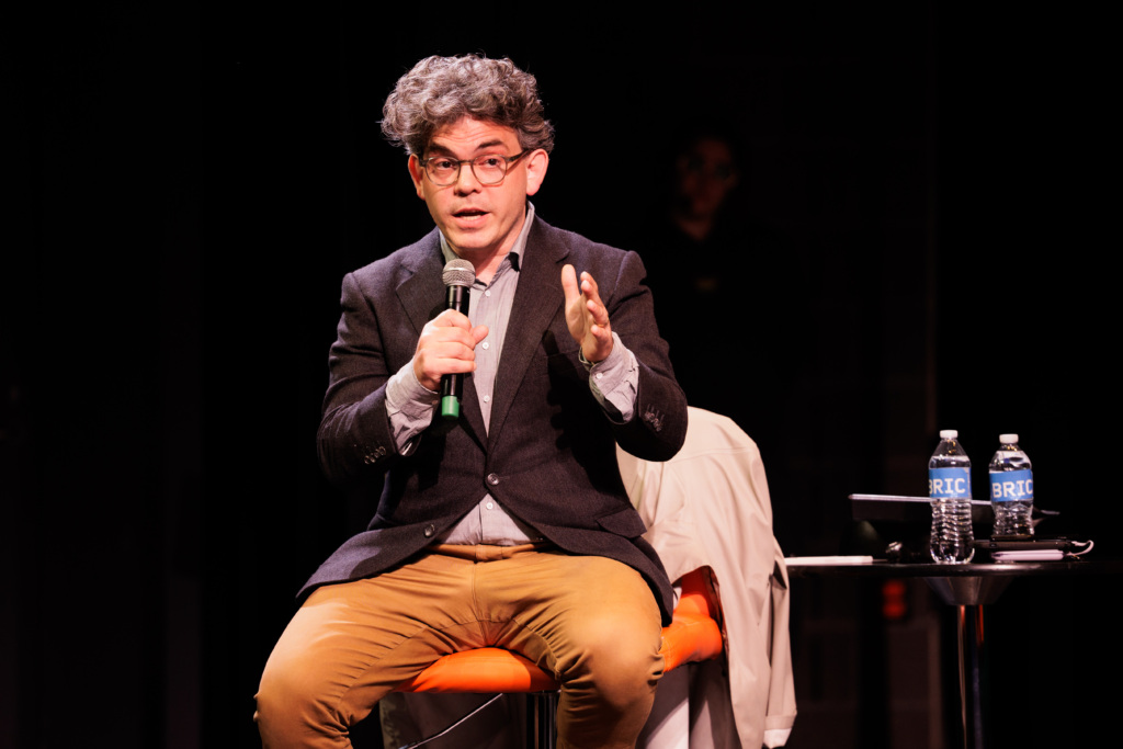 A man with glasses and curly hair sits on a stool, speaking into a microphone, gesturing with one hand. A table with two water bottles and a jacket is beside him.