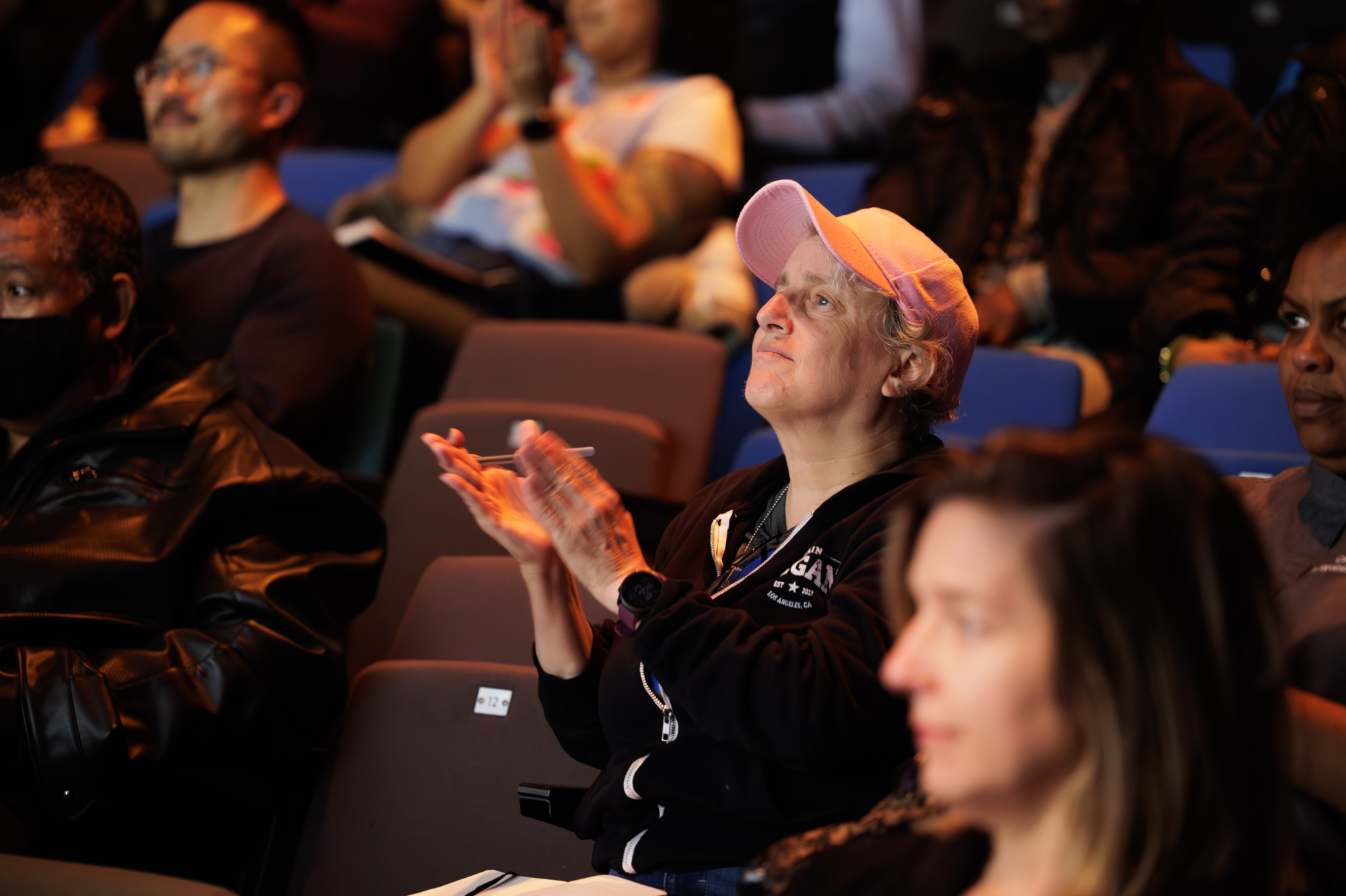 A woman in a pink cap sits in a theater audience, clapping, while others around her also watch attentively.