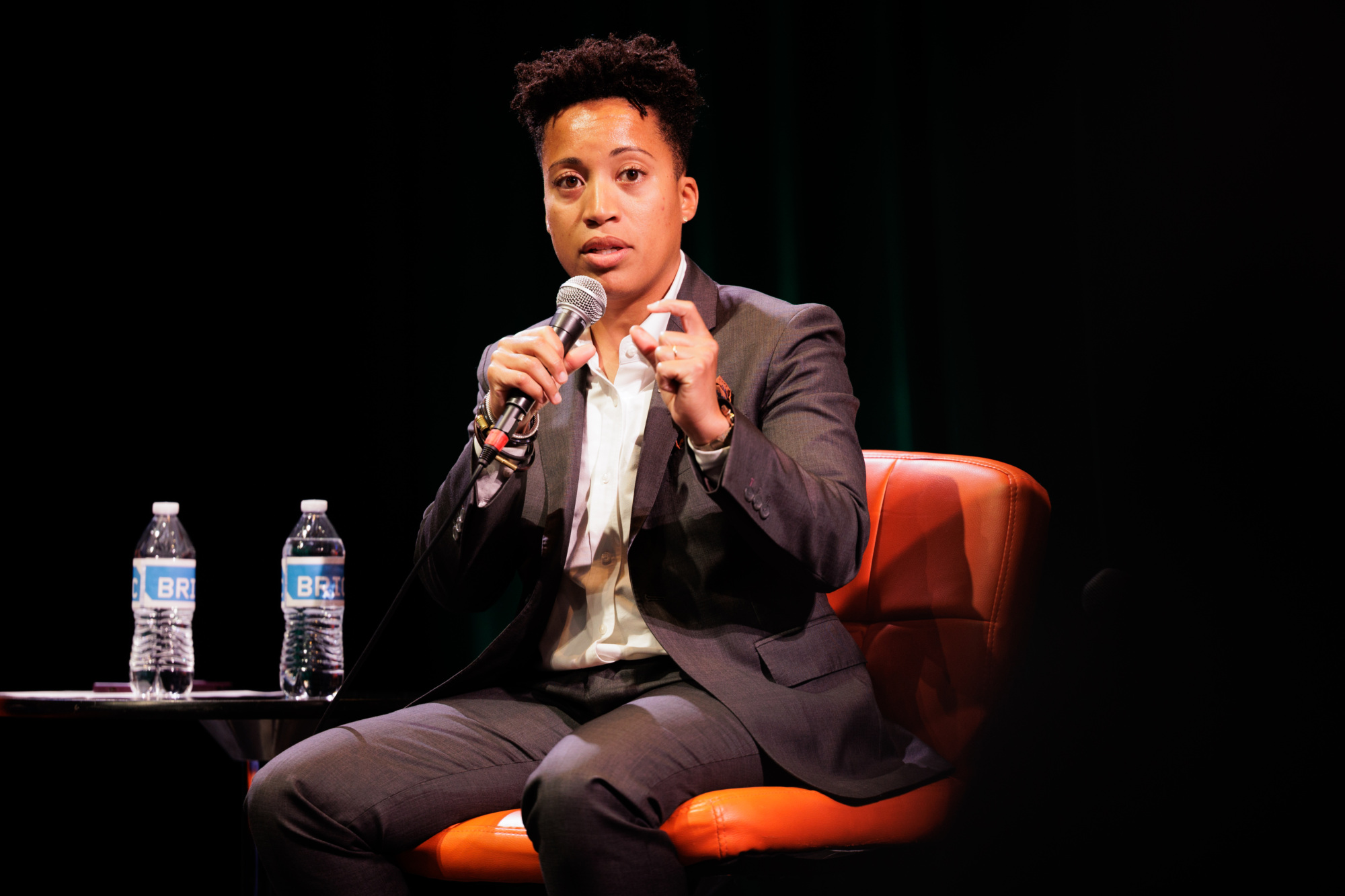 A person in a suit sits on an orange chair, speaking into a microphone on stage with two water bottles on a small table beside them.