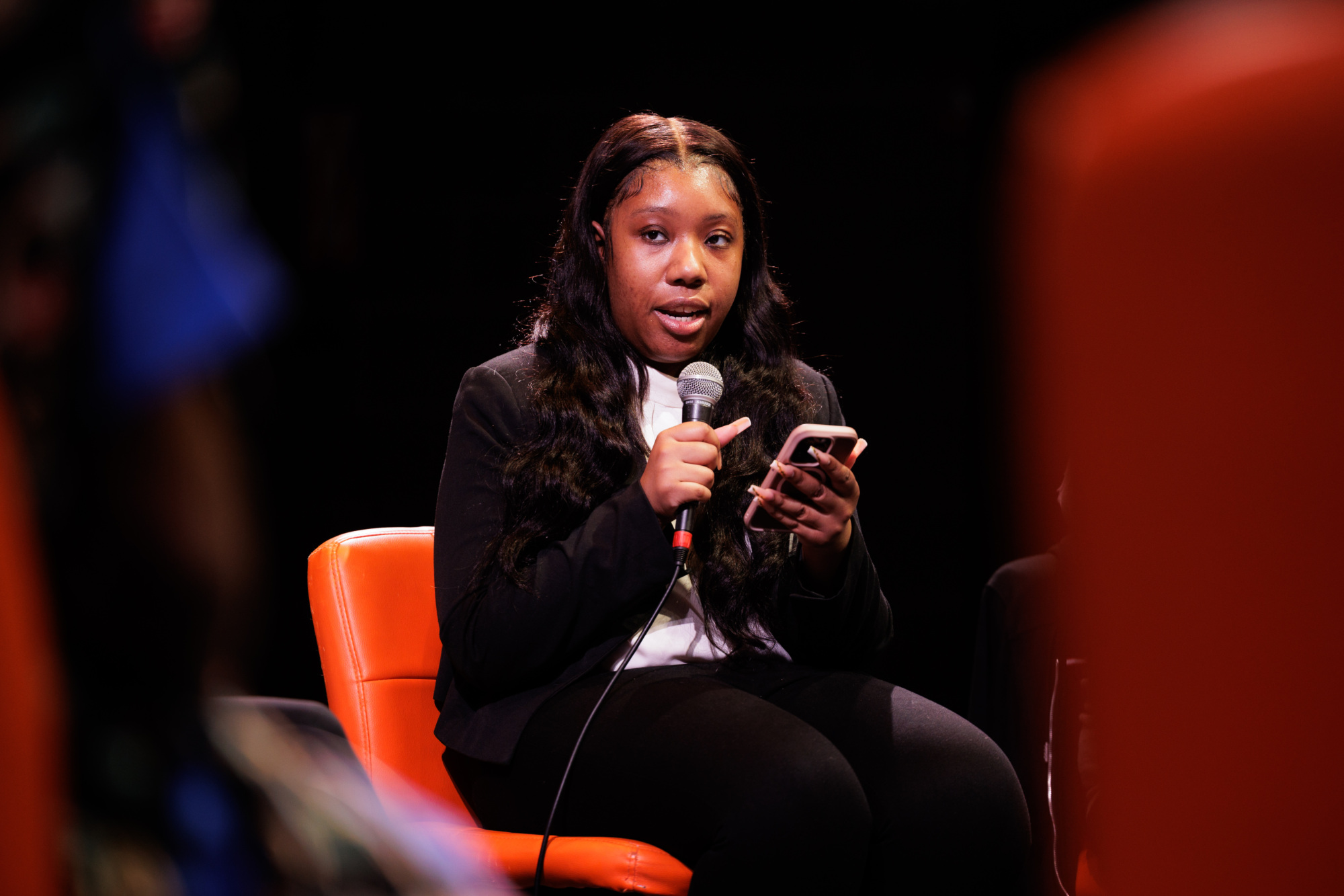 A woman sits on an orange chair, holding a microphone in one hand and a smartphone in the other, speaking during an event.