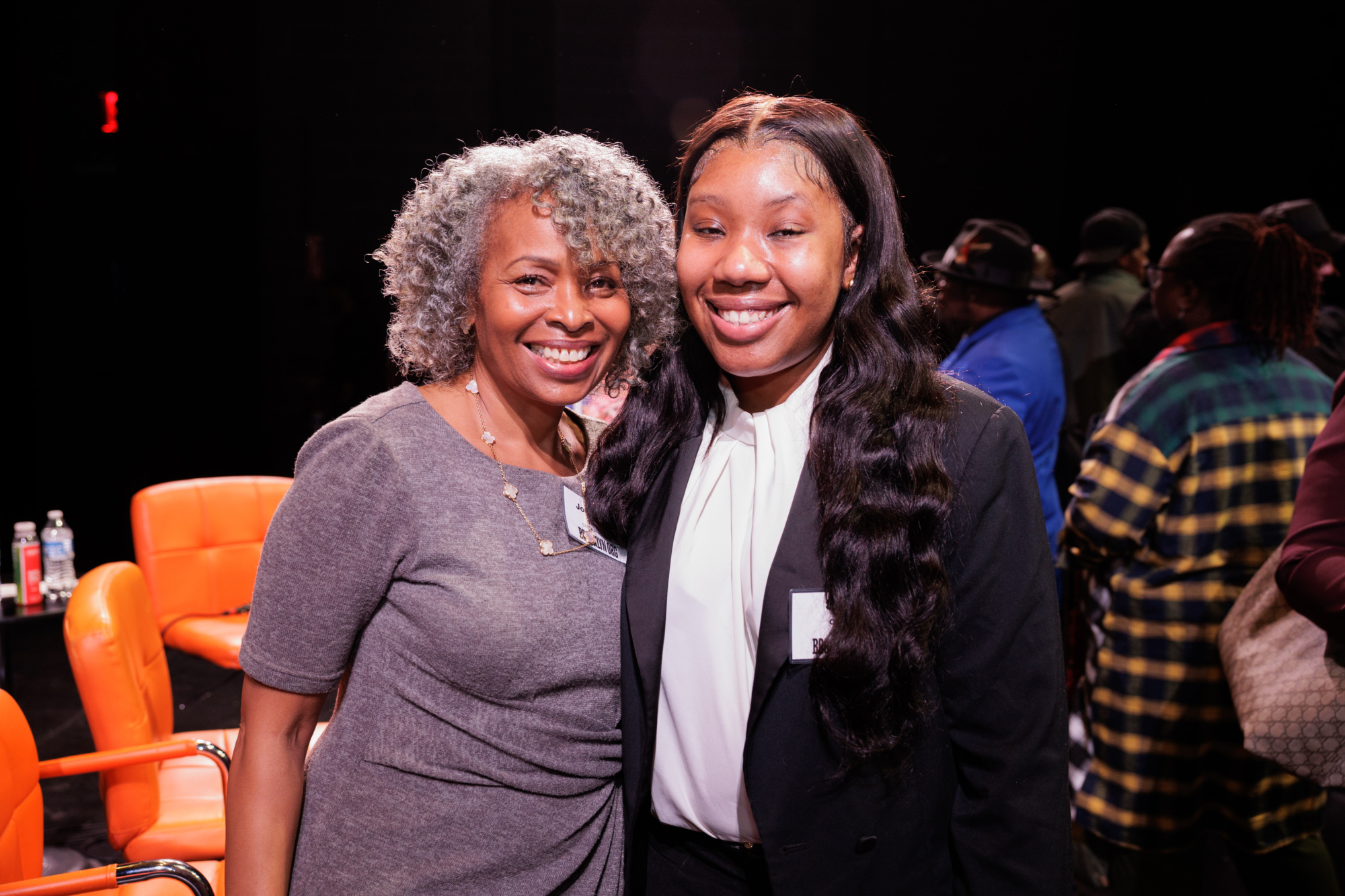 Two women stand together and smile at the camera in a room with orange chairs and other people in the background.