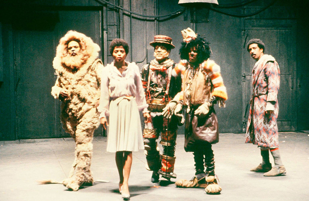 Five people in costumes stand on a stage; one is dressed as a lion, another in a white dress, and others in colorful, elaborate outfits. The background is a plain, industrial set.