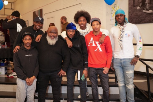 Seven men pose together indoors at an event, standing in two rows and facing the camera. Some wear hoodies and hats; balloons and event signage are visible in the background.