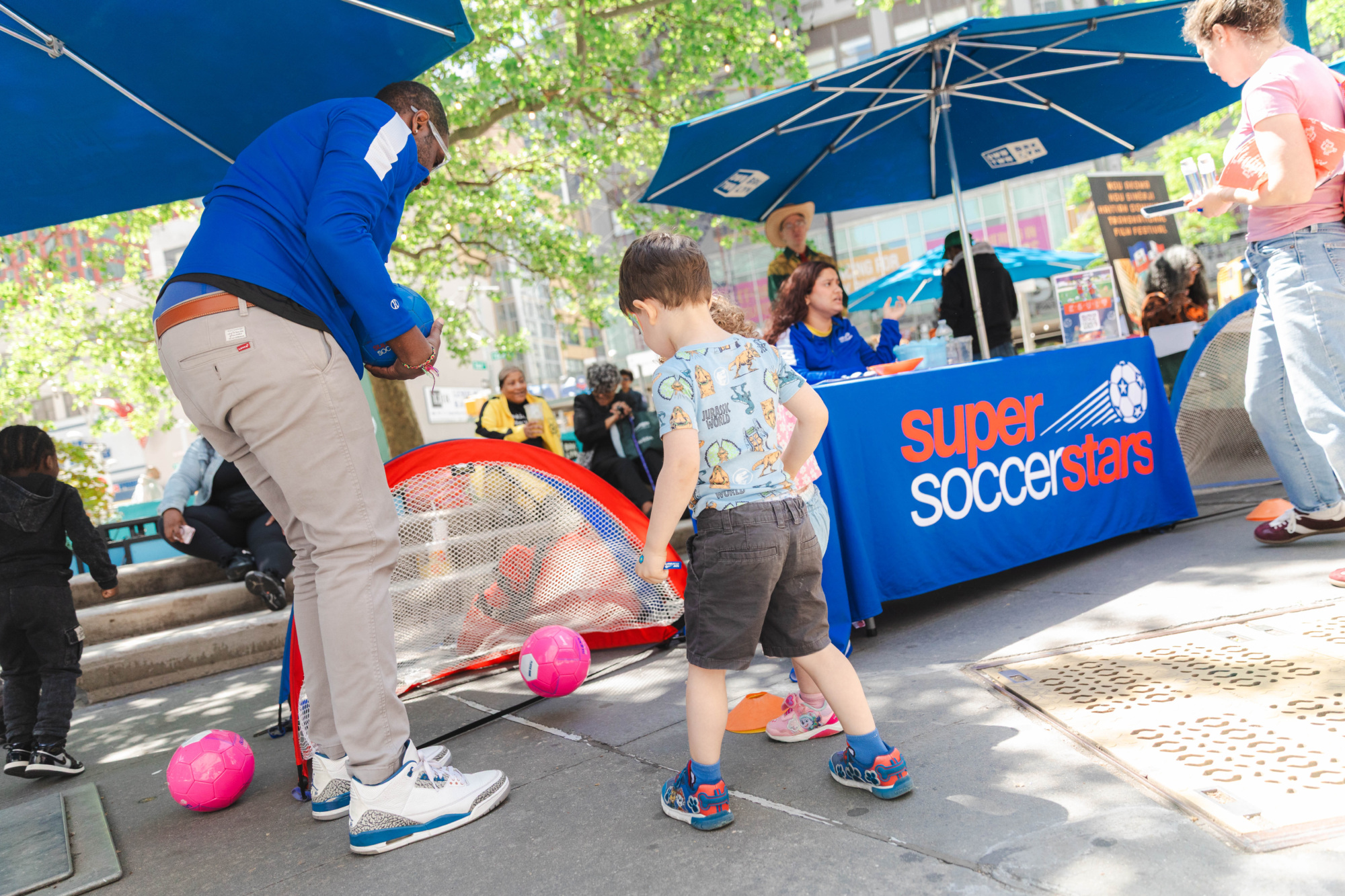 An adult helps a young child kick a soccer ball into a small net at a "Super Soccer Stars" outdoor event, with people seated at a nearby table under umbrellas.