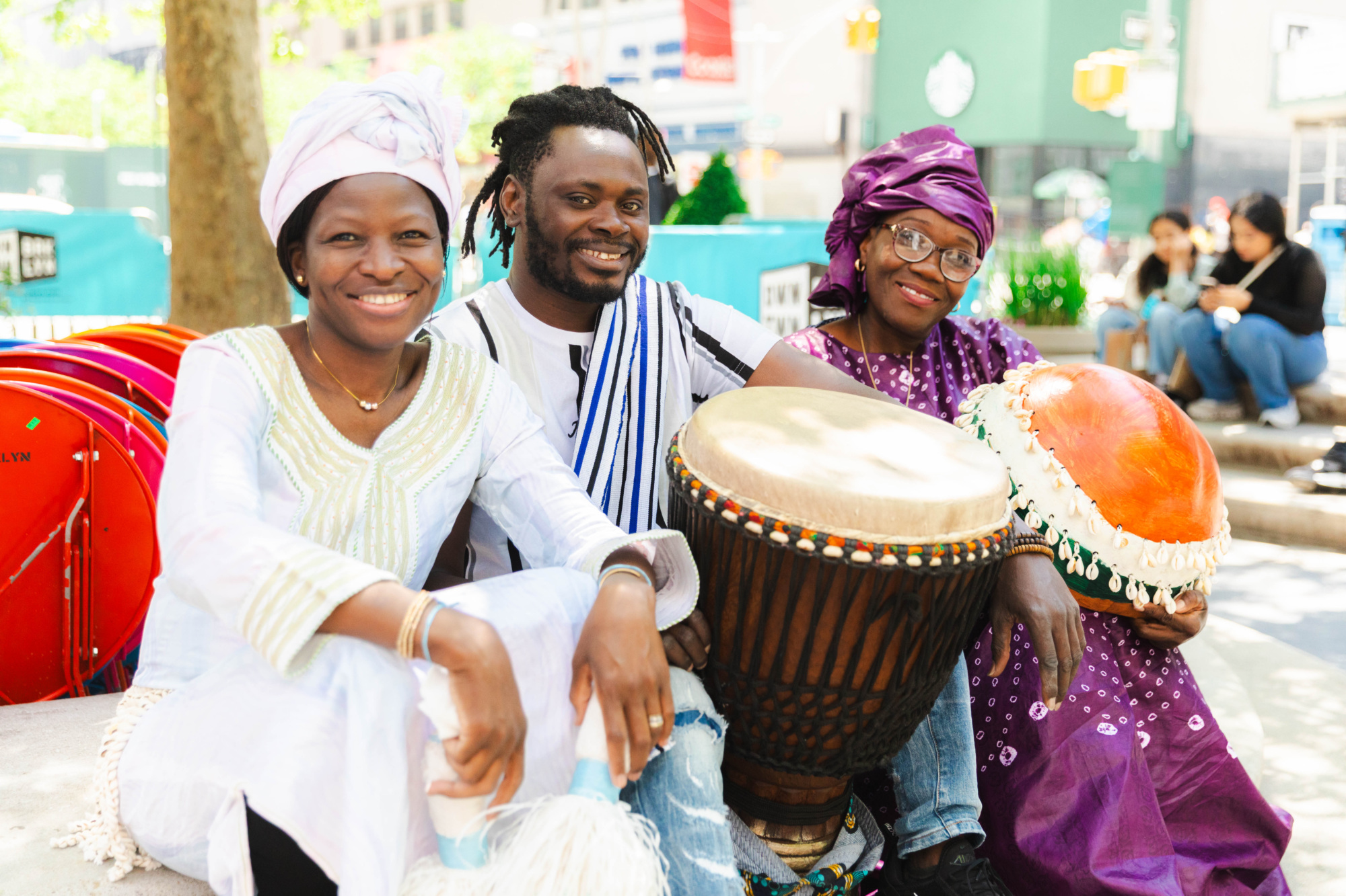 Three people in traditional clothing sit outdoors, smiling, holding large hand drums and percussion instruments in a public urban setting.