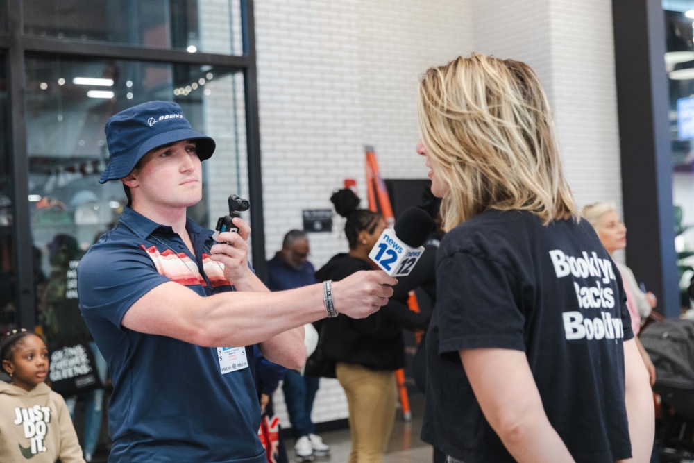 A reporter wearing a blue hat interviews a woman in a "Booklyn Tactics Bodhi" T-shirt indoors; people are gathered in the background.