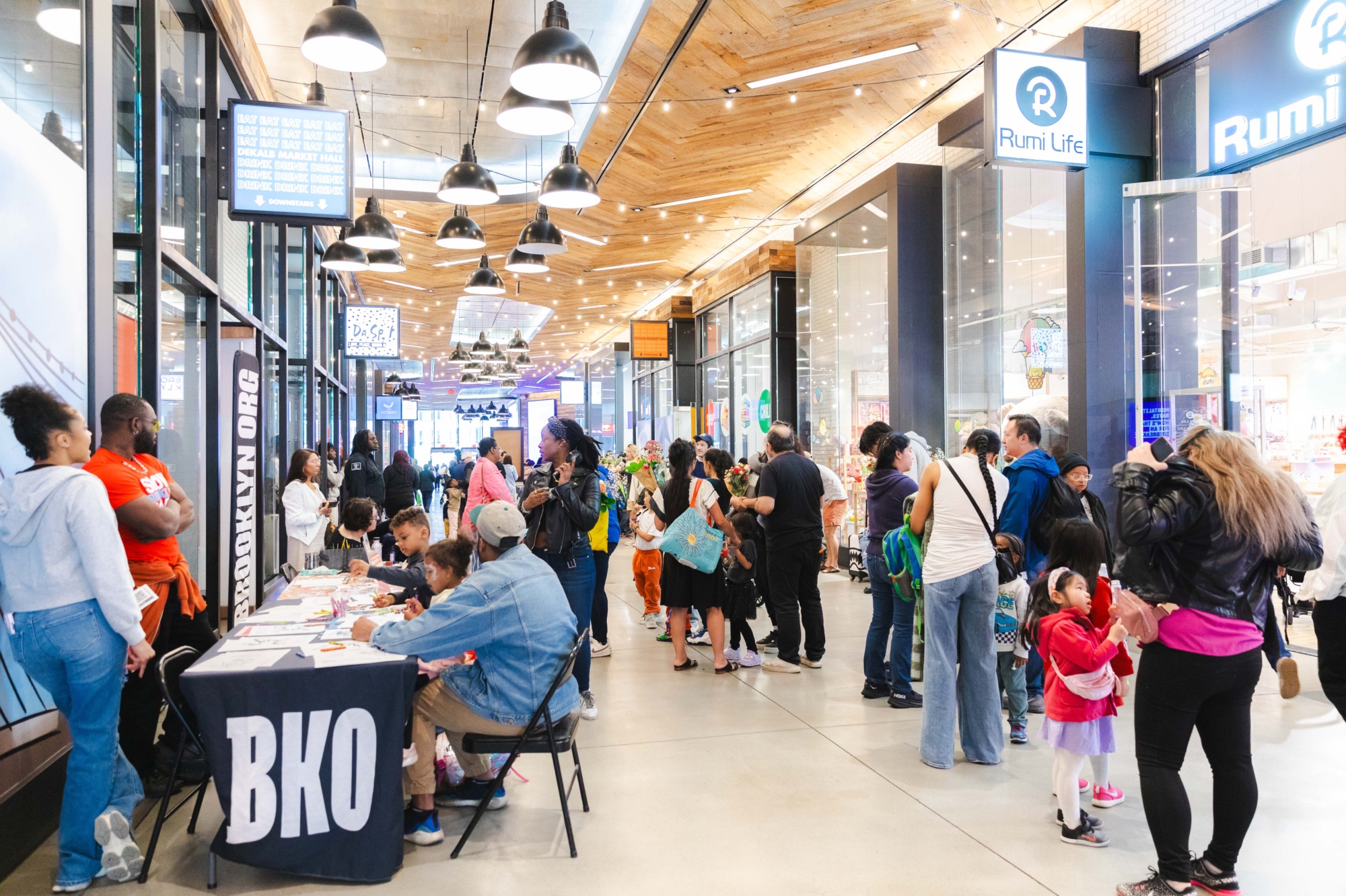 A busy indoor mall hallway with people lined up and gathered at tables with informational signs; children and adults interact near shop entrances.