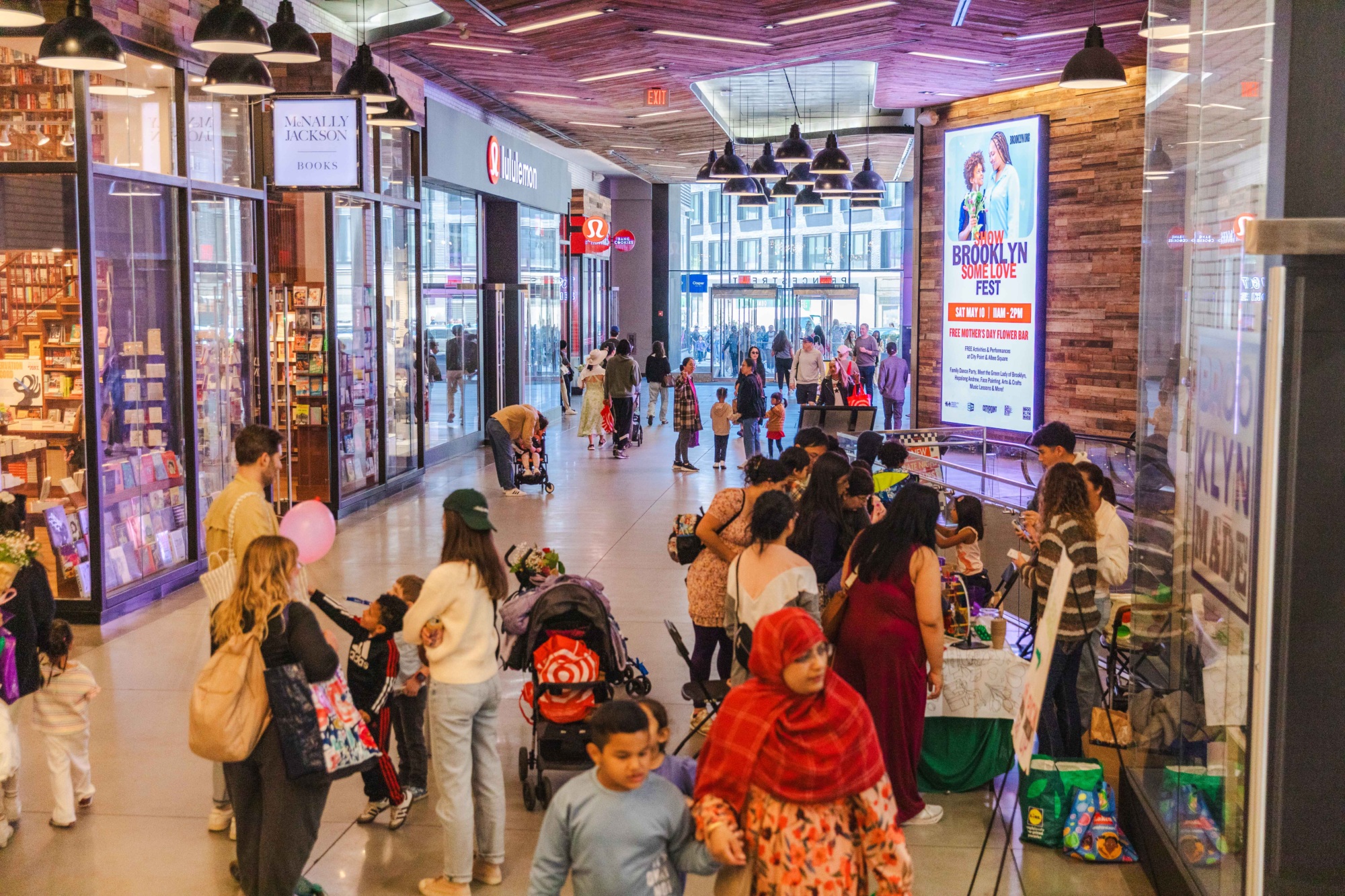 A busy shopping mall corridor with people walking, some pushing strollers, and groups gathered near stores and a large digital advertisement.