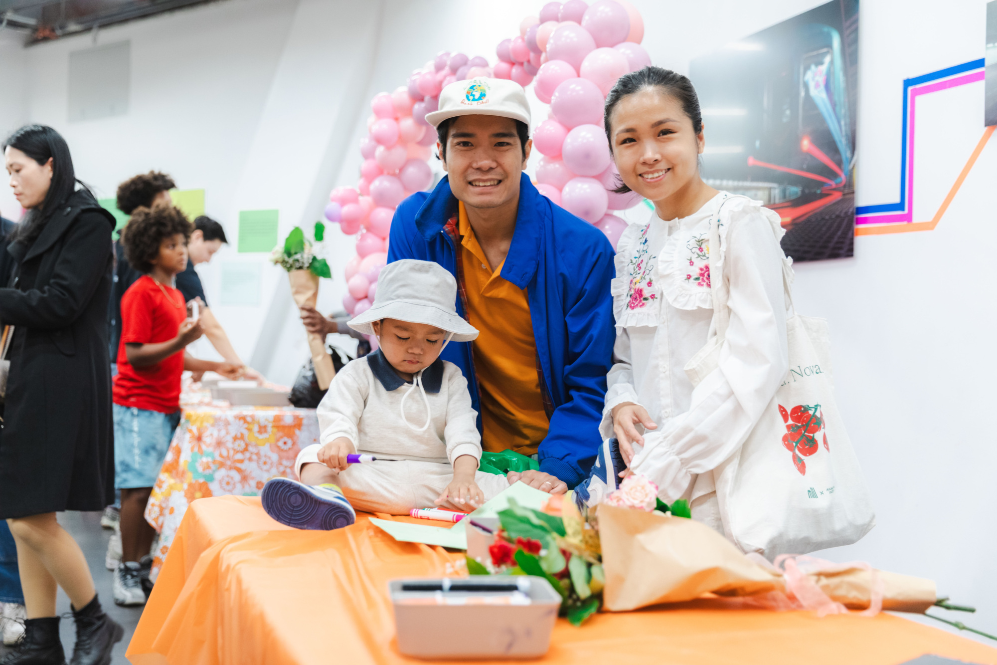 A family of three poses at a decorated table with flowers and art supplies, while people gather in the background at an indoor event with pink balloons.