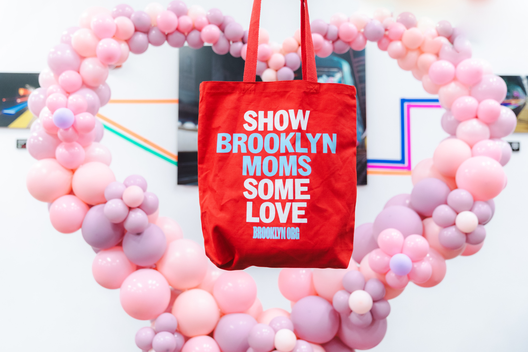 A red tote bag with the words "Show Brooklyn Moms Some Love" hangs in front of a pink and purple balloon heart display.