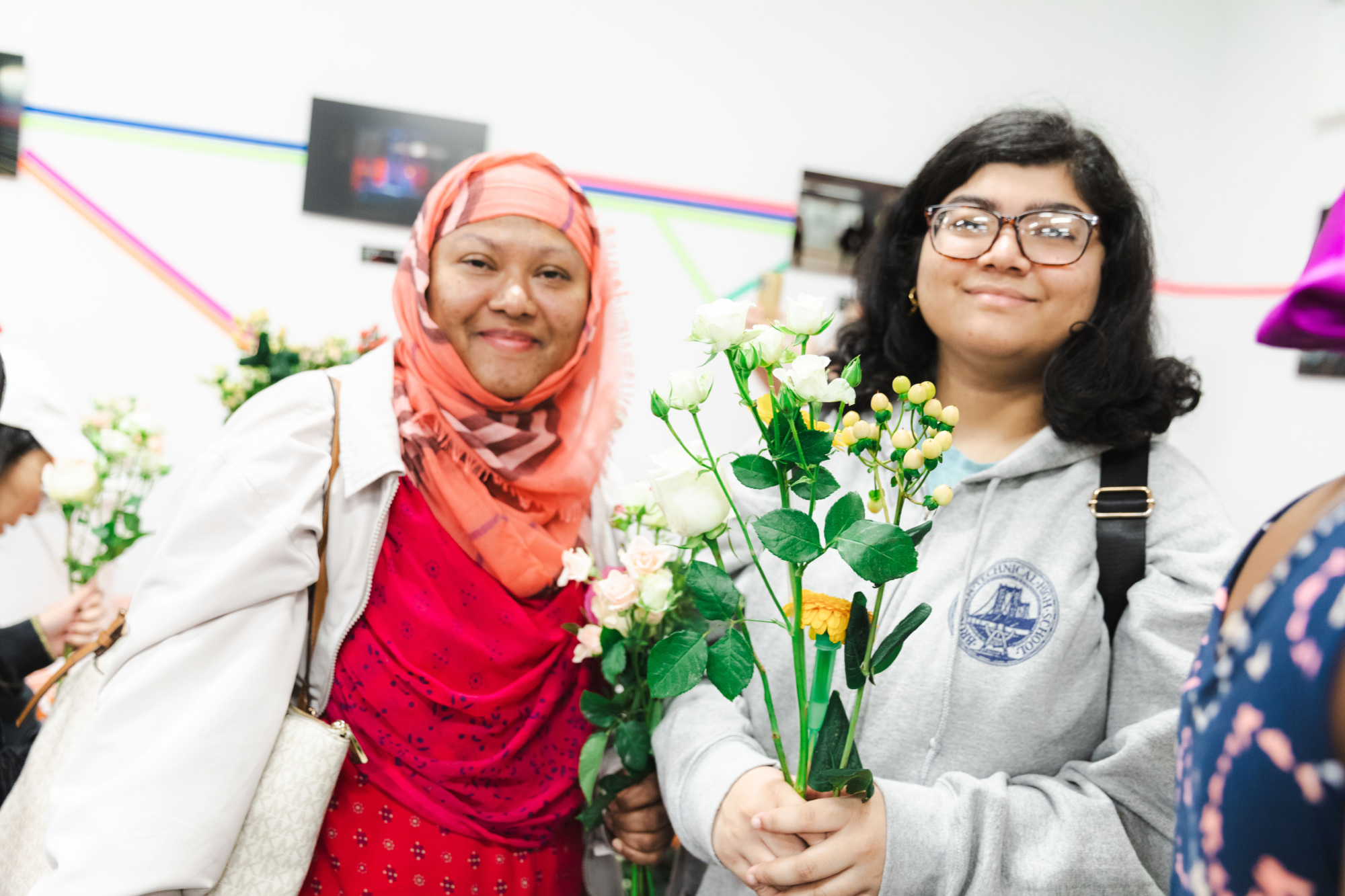 Two women stand indoors holding bouquets of flowers, smiling at the camera, with colorful lines and framed photos on the wall behind them.