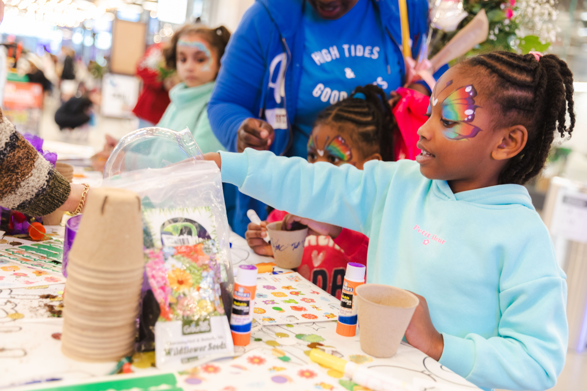 A child with butterfly face paint reaches across a table with craft supplies, surrounded by other children and adults at an indoor event.