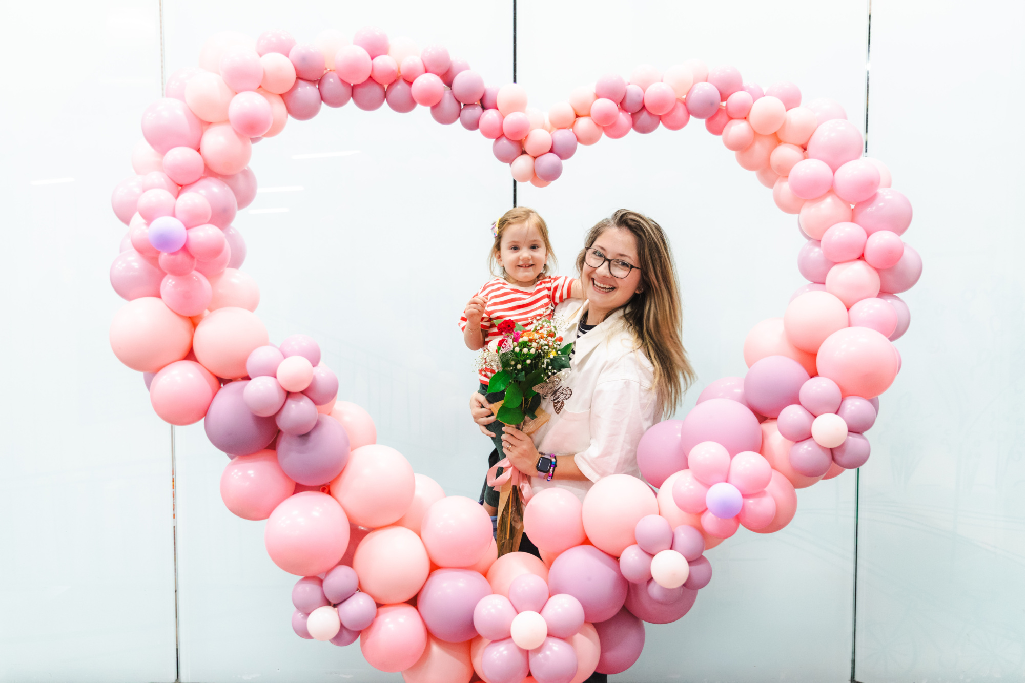 An adult and a child stand together holding flowers inside a heart-shaped balloon frame made of pink and purple balloons against a white background.