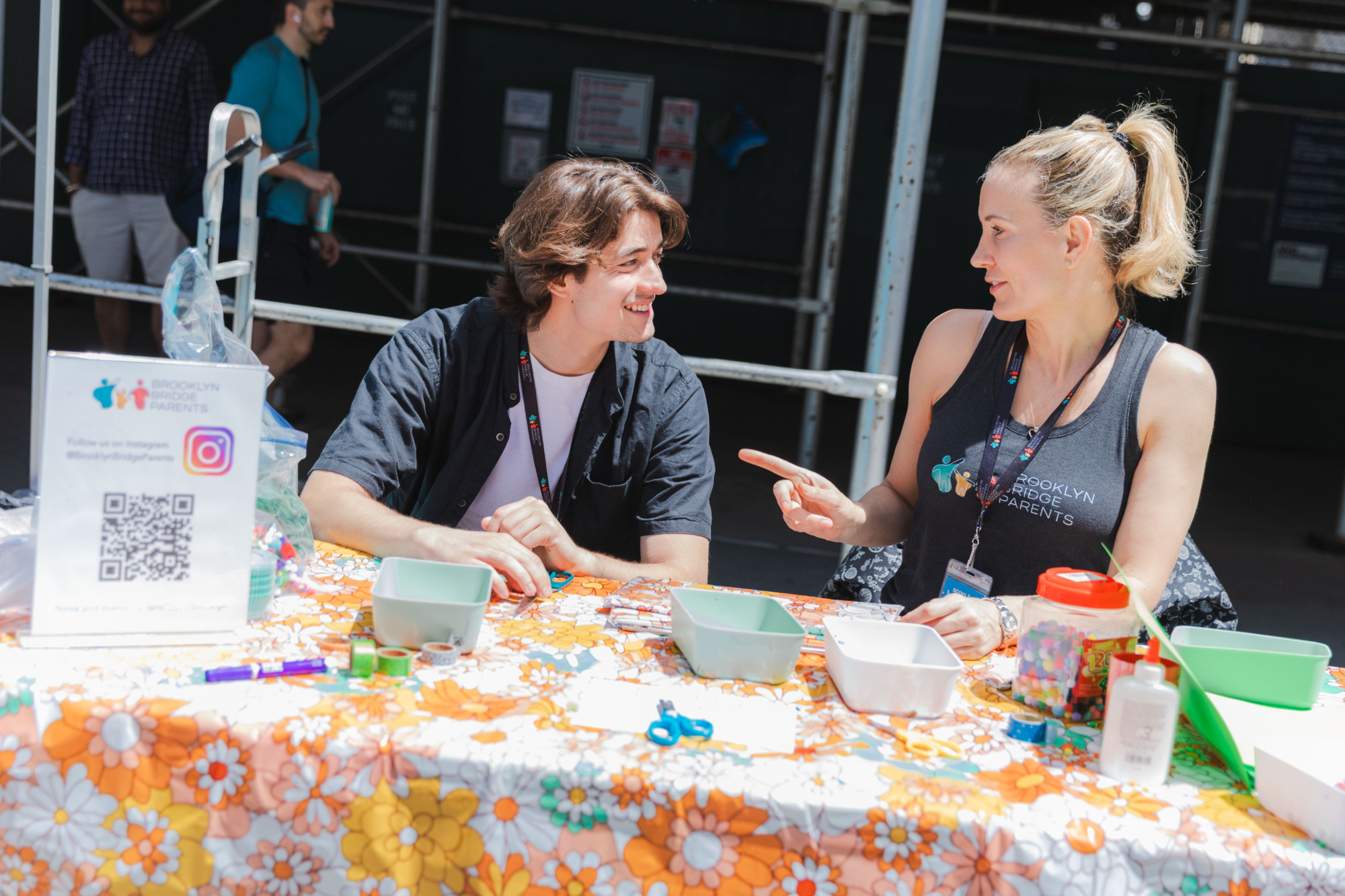 Two people sit at a flower-patterned table outdoors, talking. Craft supplies, a QR code sign, and containers are on the table. A third person stands in the background.