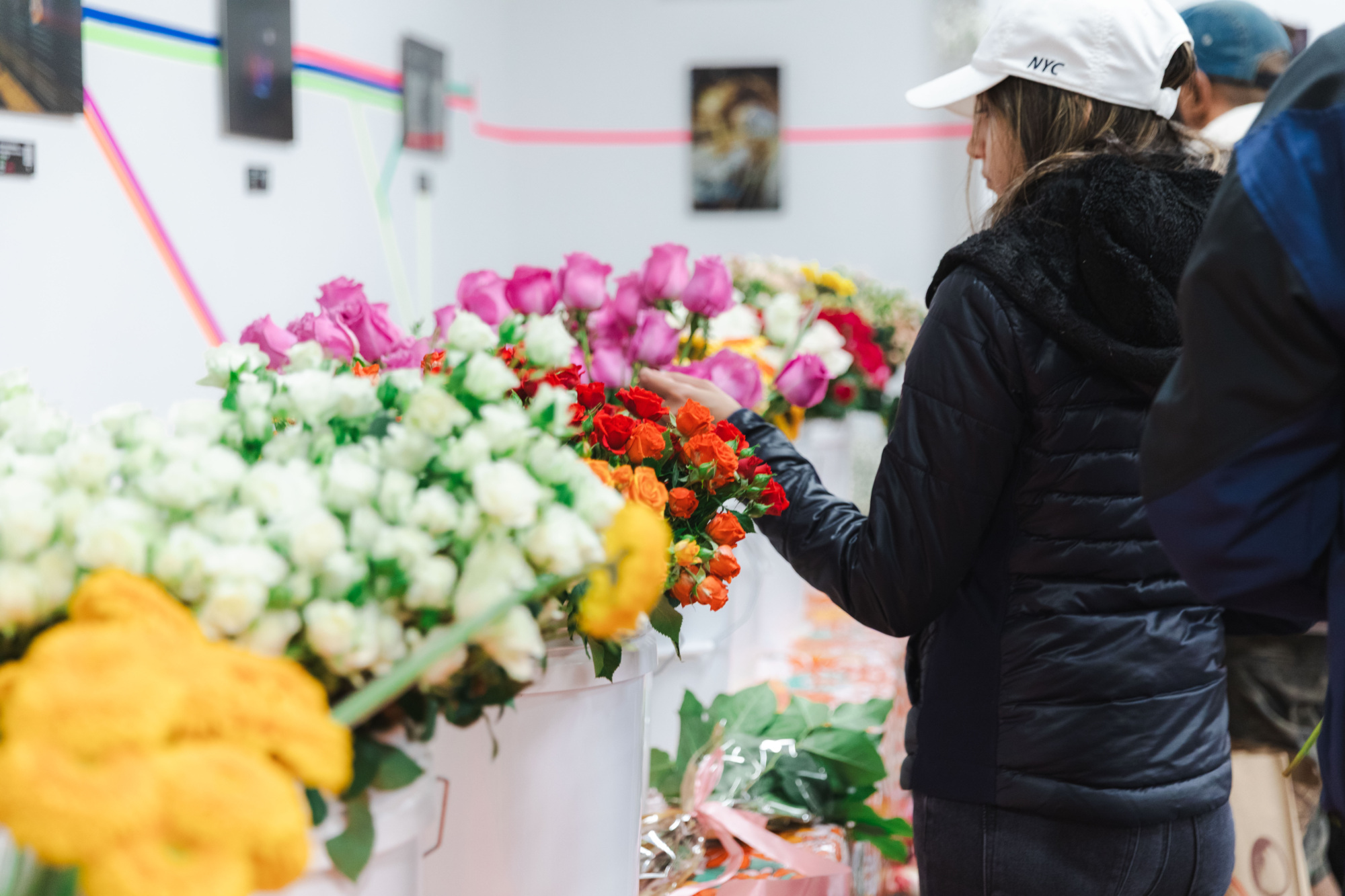 A person wearing a white cap and black jacket selects flowers from several colorful bouquets displayed on a table indoors.