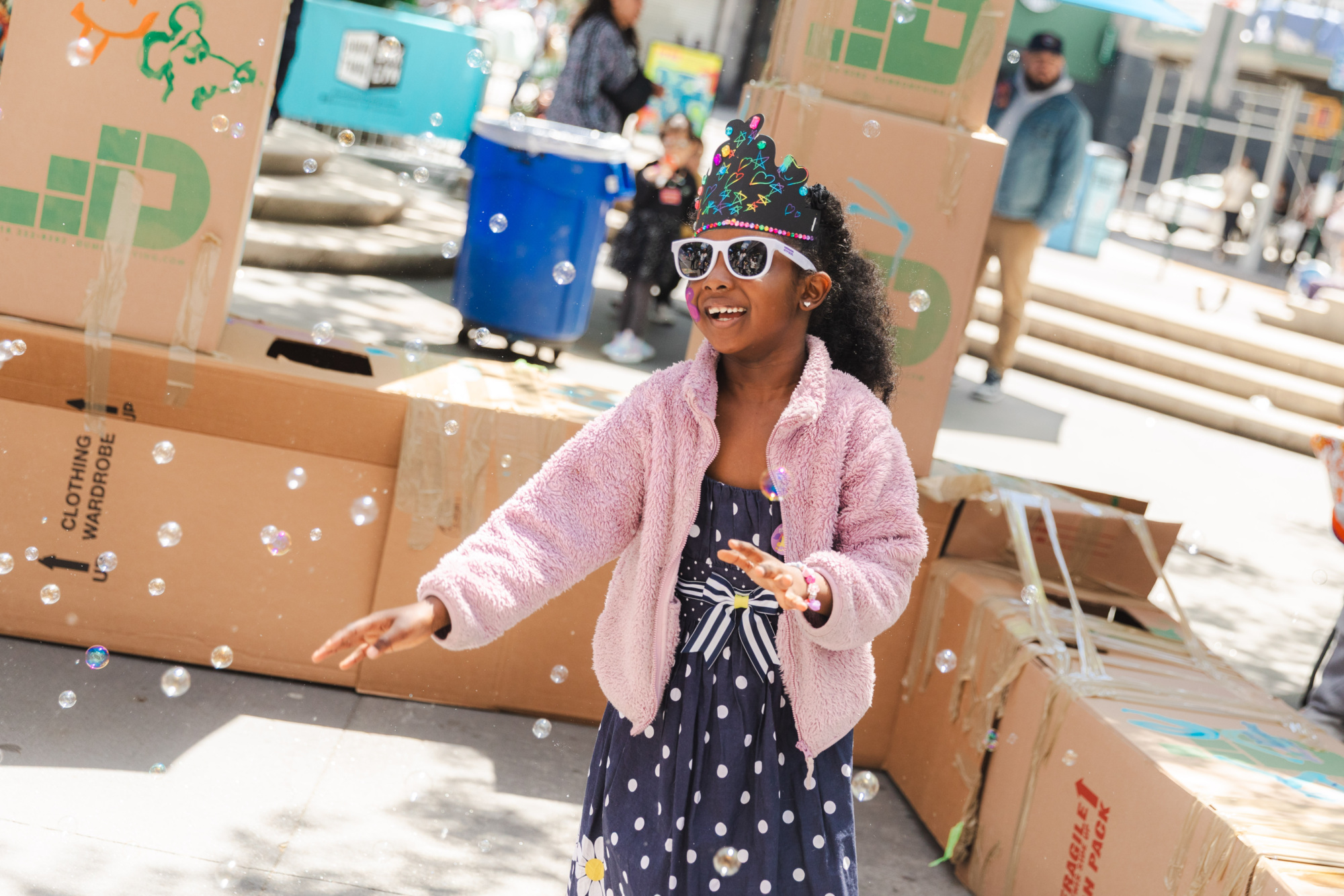 A girl in a polka dot dress and crown smiles and reaches for bubbles while standing in front of stacked cardboard boxes outdoors.