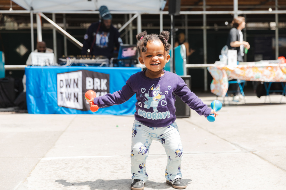 A young child smiles while holding maracas, standing outdoors in front of a DJ booth at a street event.