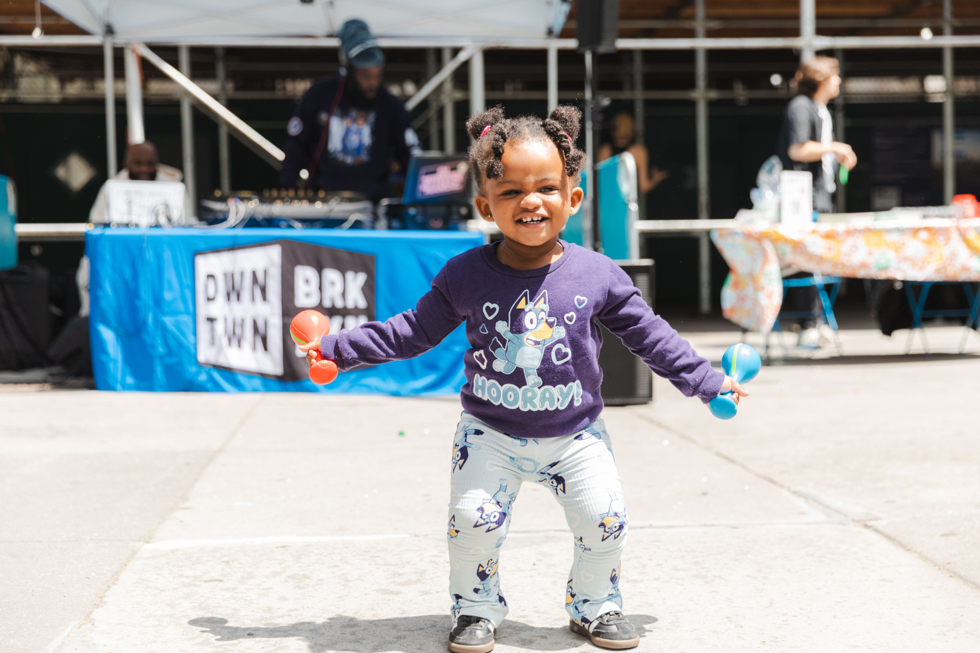 A young child smiles while holding maracas, standing outdoors in front of a DJ booth at a street event.