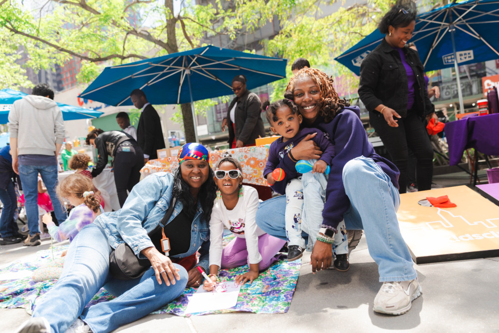 Three adults and a child sit and pose for a photo at an outdoor event, surrounded by other people, under blue umbrellas and trees.