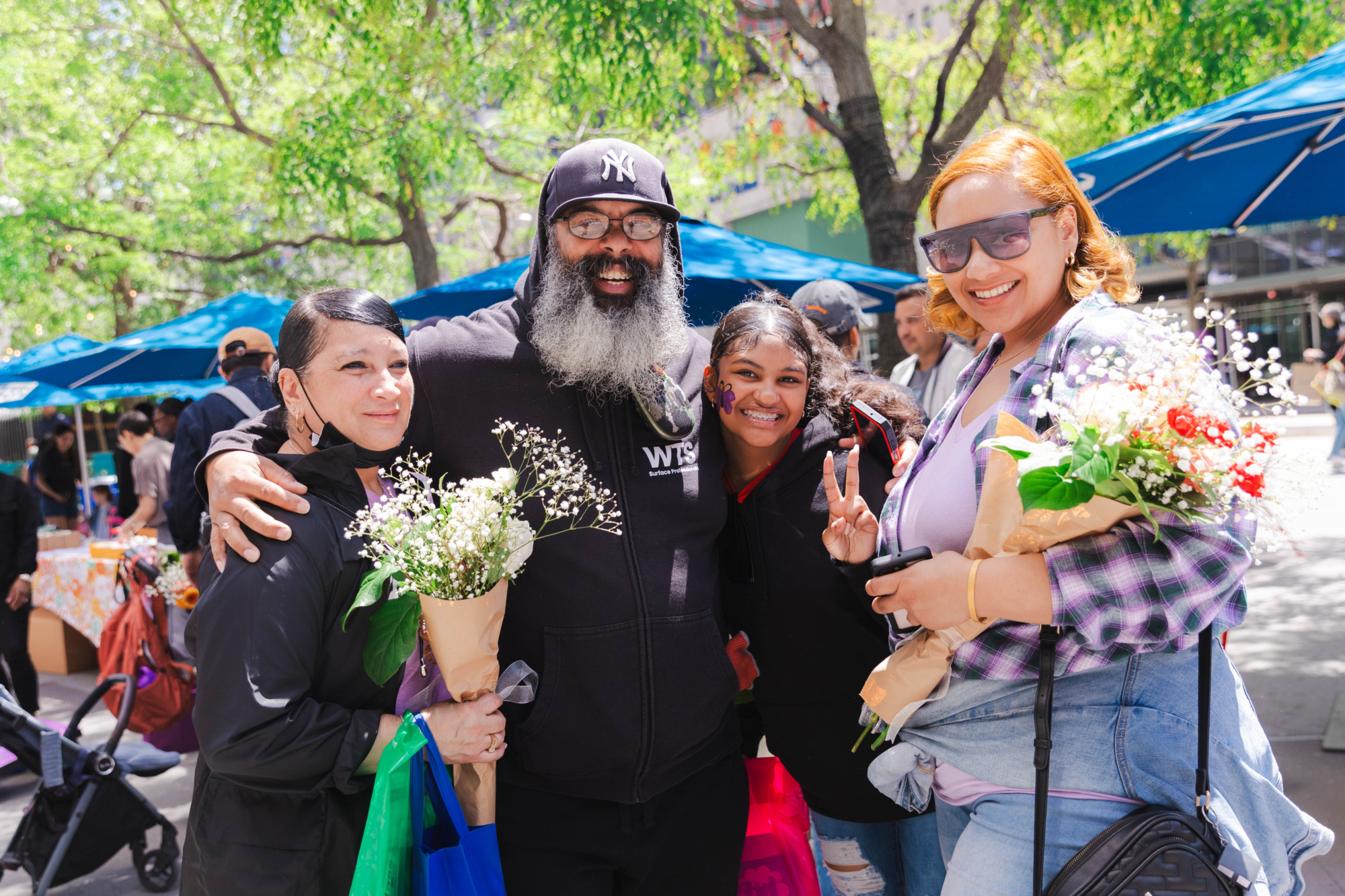 Four people stand outdoors smiling, two holding flower bouquets, with trees and blue umbrellas in the background.