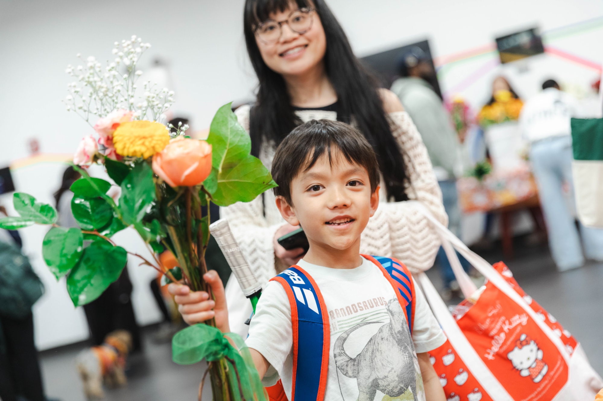 A young boy holding flowers stands in front of an adult woman, both smiling at the camera in a busy indoor setting.