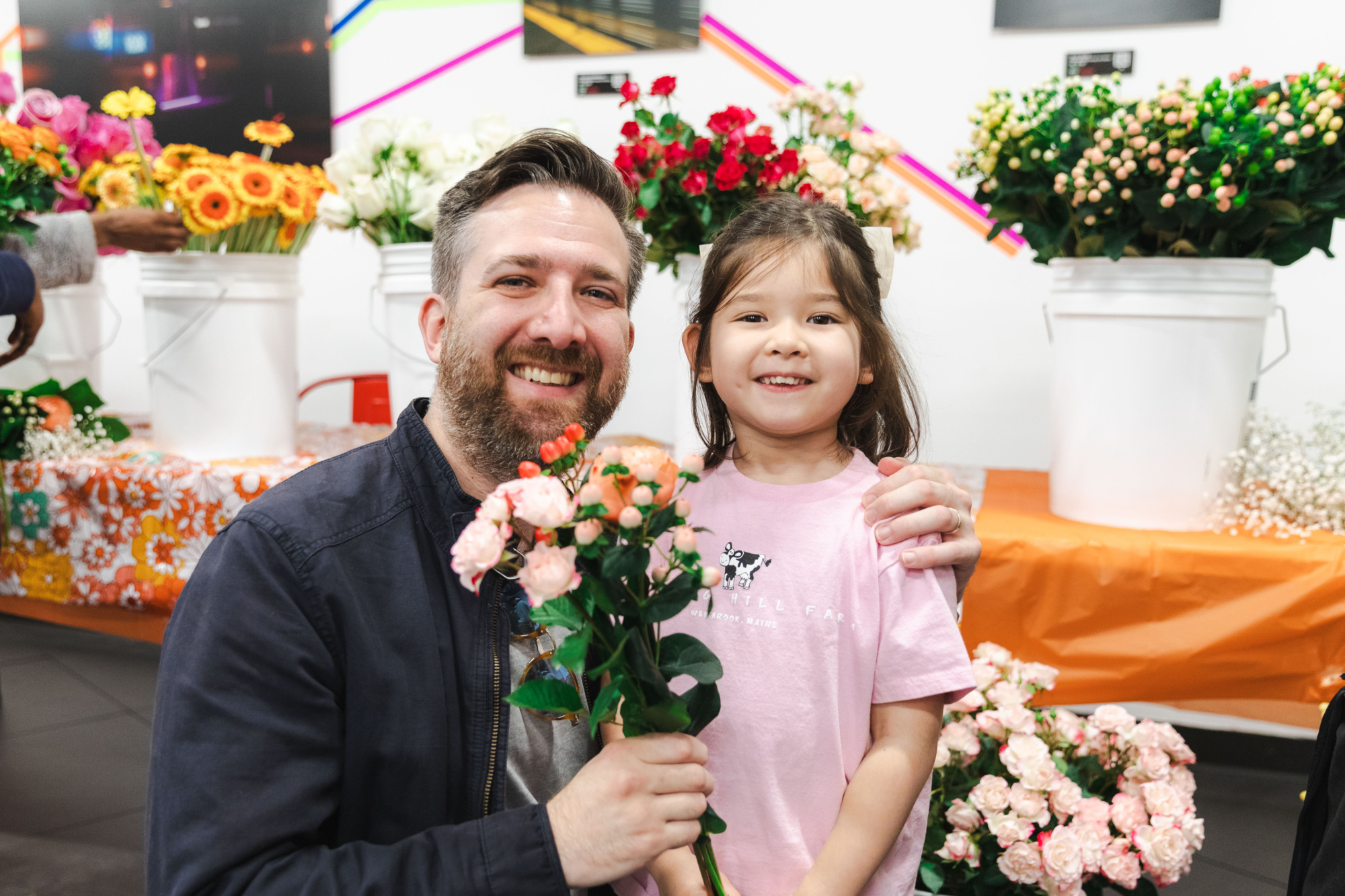 A man and a young girl smile while holding flowers in front of buckets of assorted blooms at an indoor market or flower shop.
