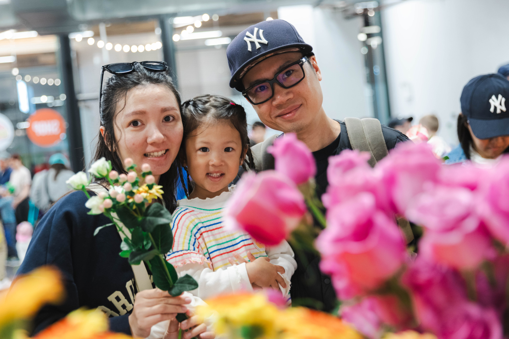 A woman, man, and young girl stand together holding flowers, smiling at the camera in a market setting with bright pink and yellow flowers in the foreground.