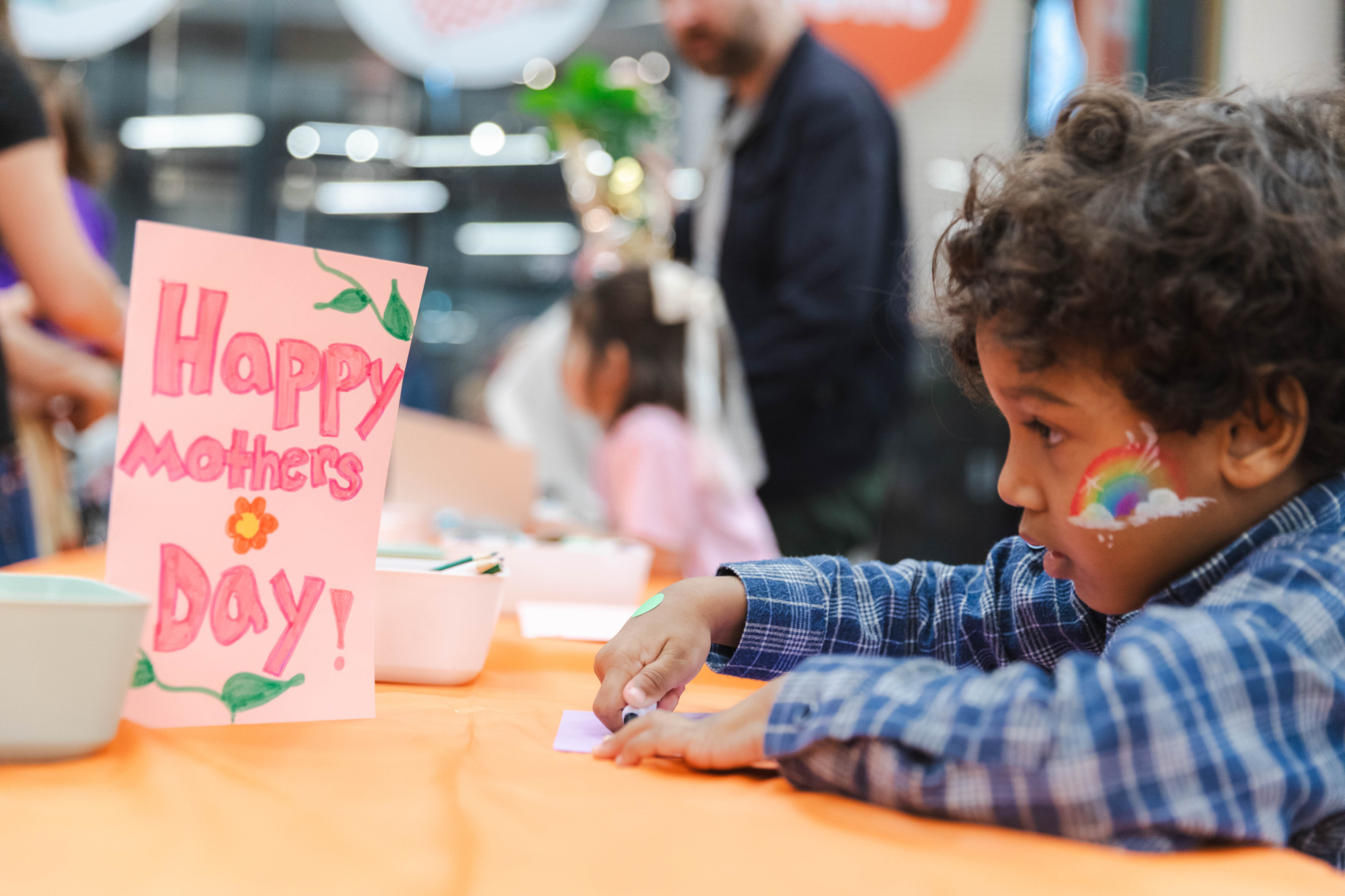 A young child with a rainbow painted on their face sits at a table making crafts next to a handmade "Happy Mother's Day!" card.