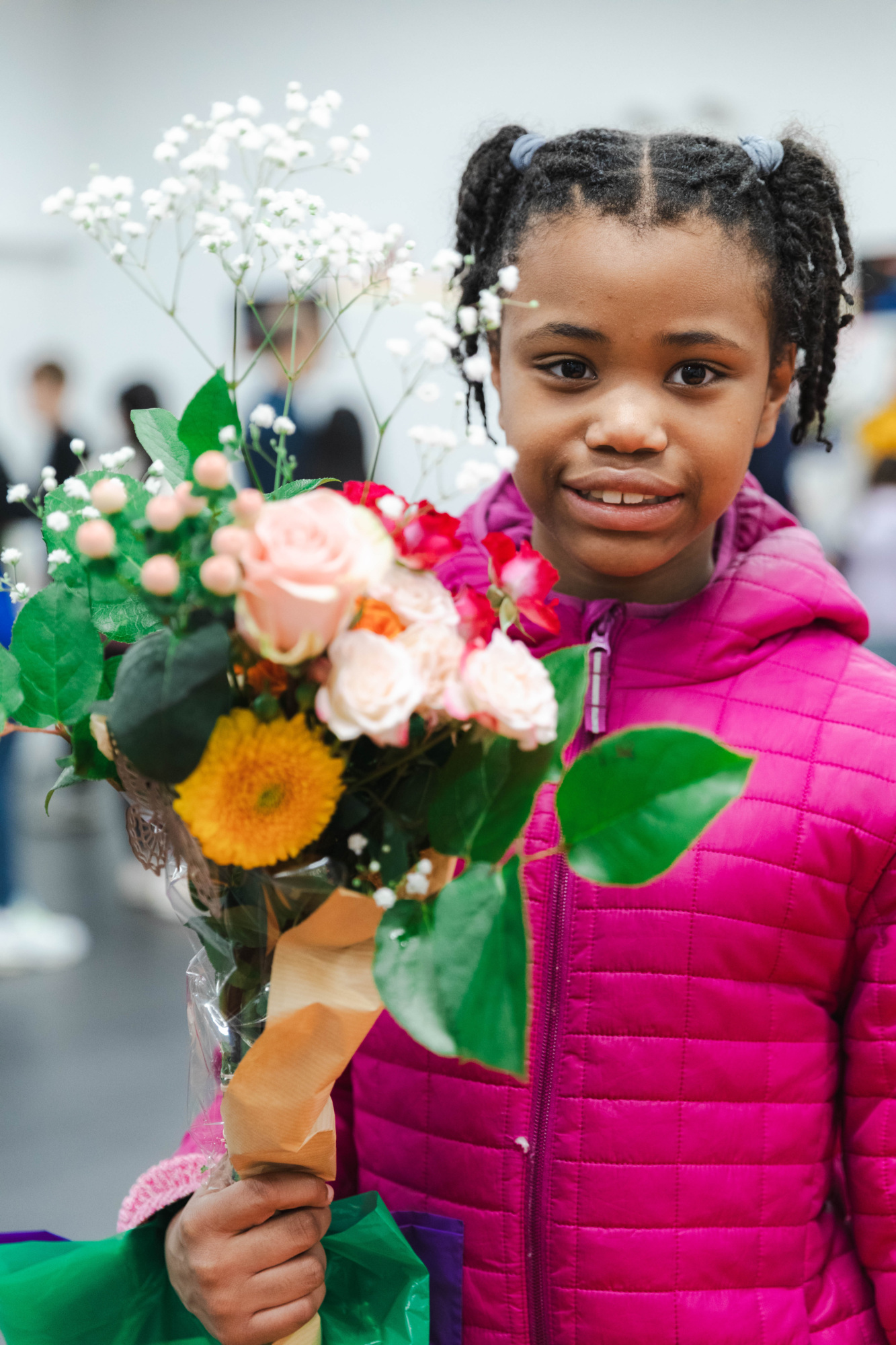 A young girl in a pink jacket holds a colorful bouquet of flowers and smiles at the camera.
