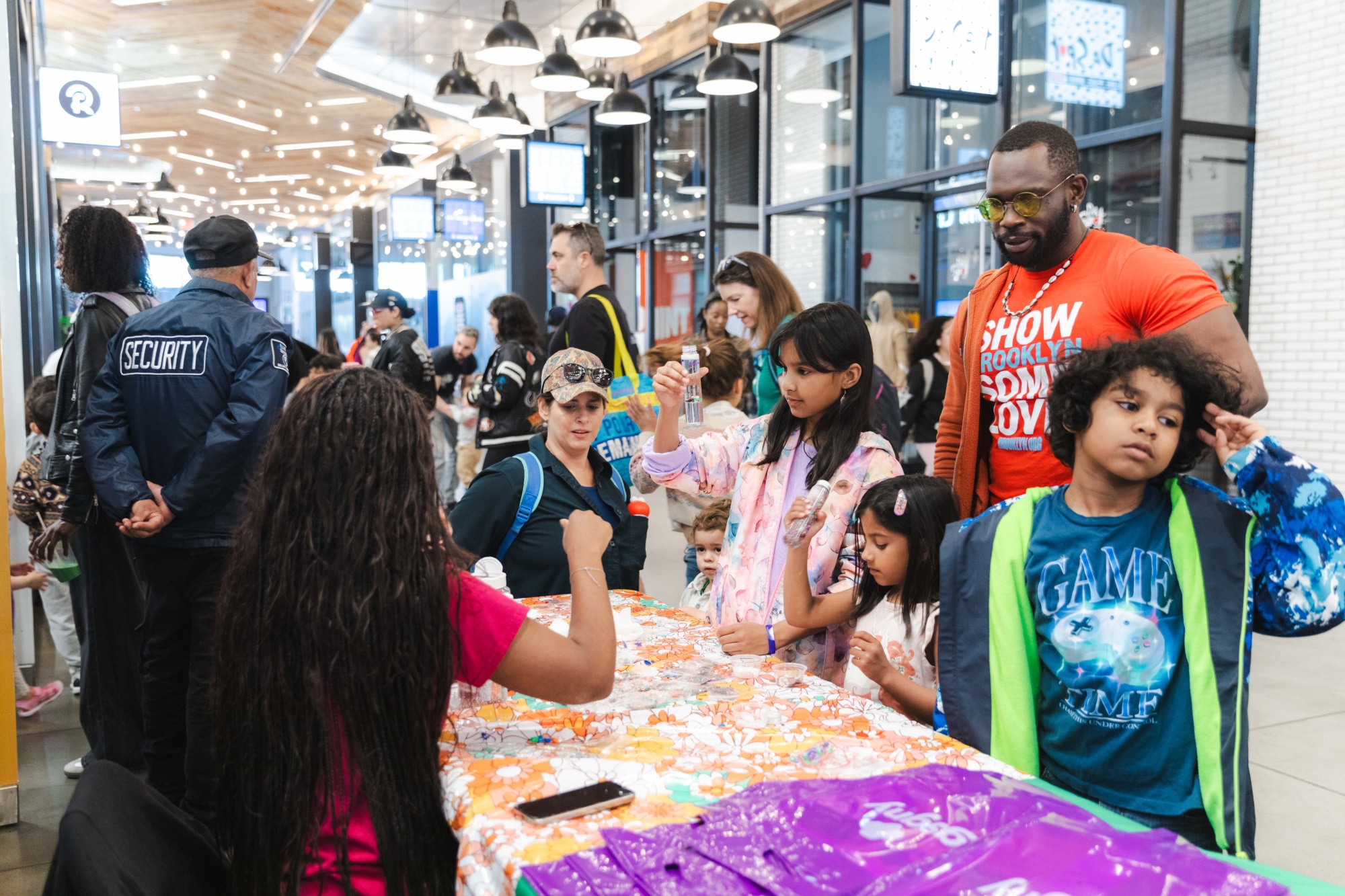 Children and adults gather around a table with crafts and activities at a busy indoor event, with security personnel and bystanders in the background.