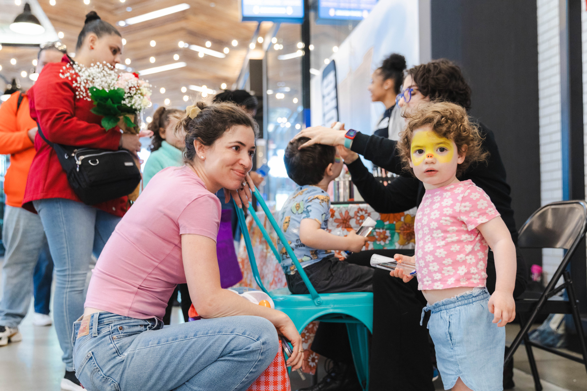 A child with yellow face paint stands next to an adult, while other children and adults sit or stand nearby at an indoor public event.