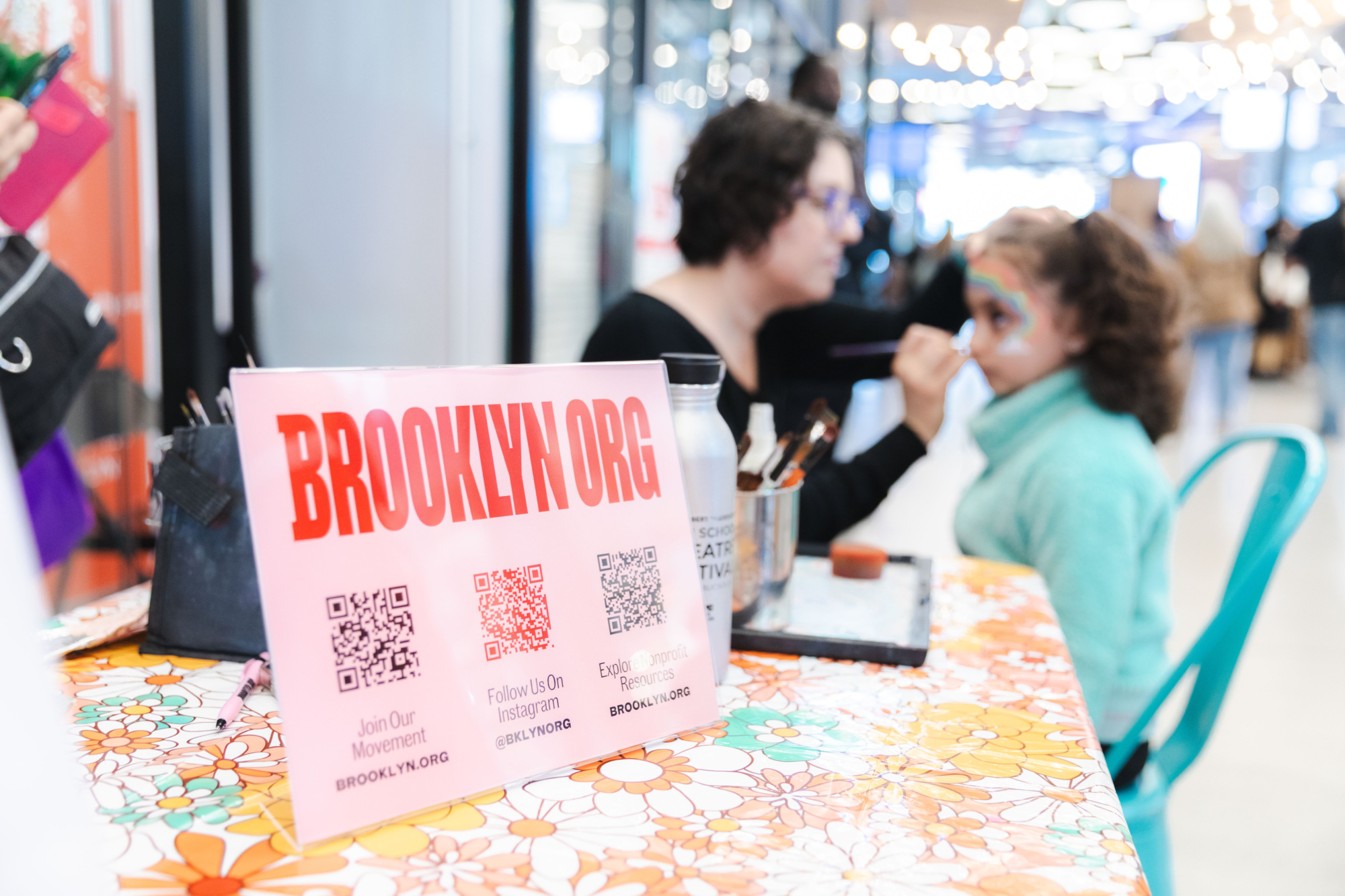 A woman paints a child's face at a table with a floral tablecloth; a pink BROOKLYN.ORG sign with QR codes is displayed in the foreground.