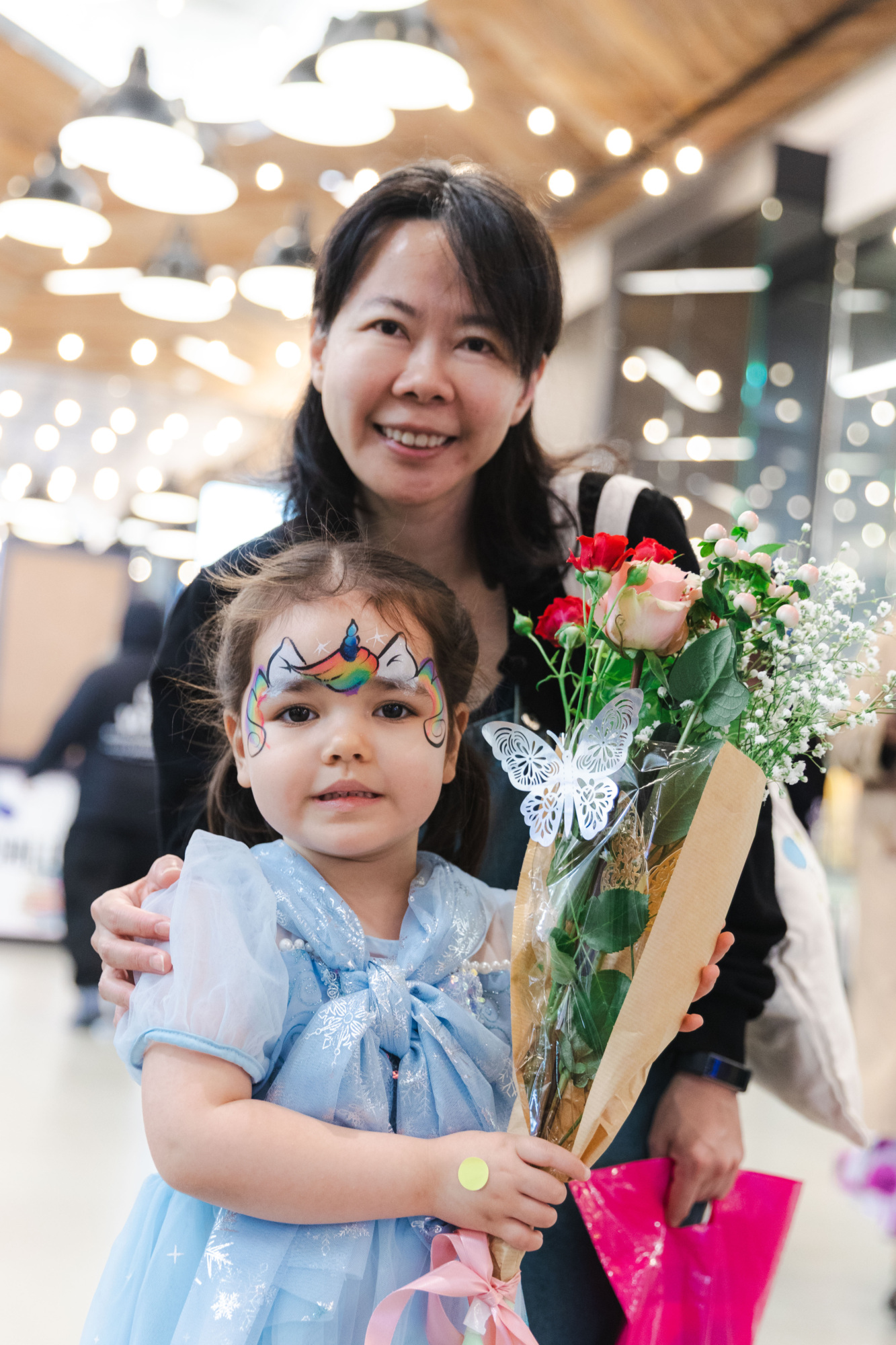 A young girl in a blue costume with face paint holds a bouquet of flowers, standing in front of an adult woman indoors.