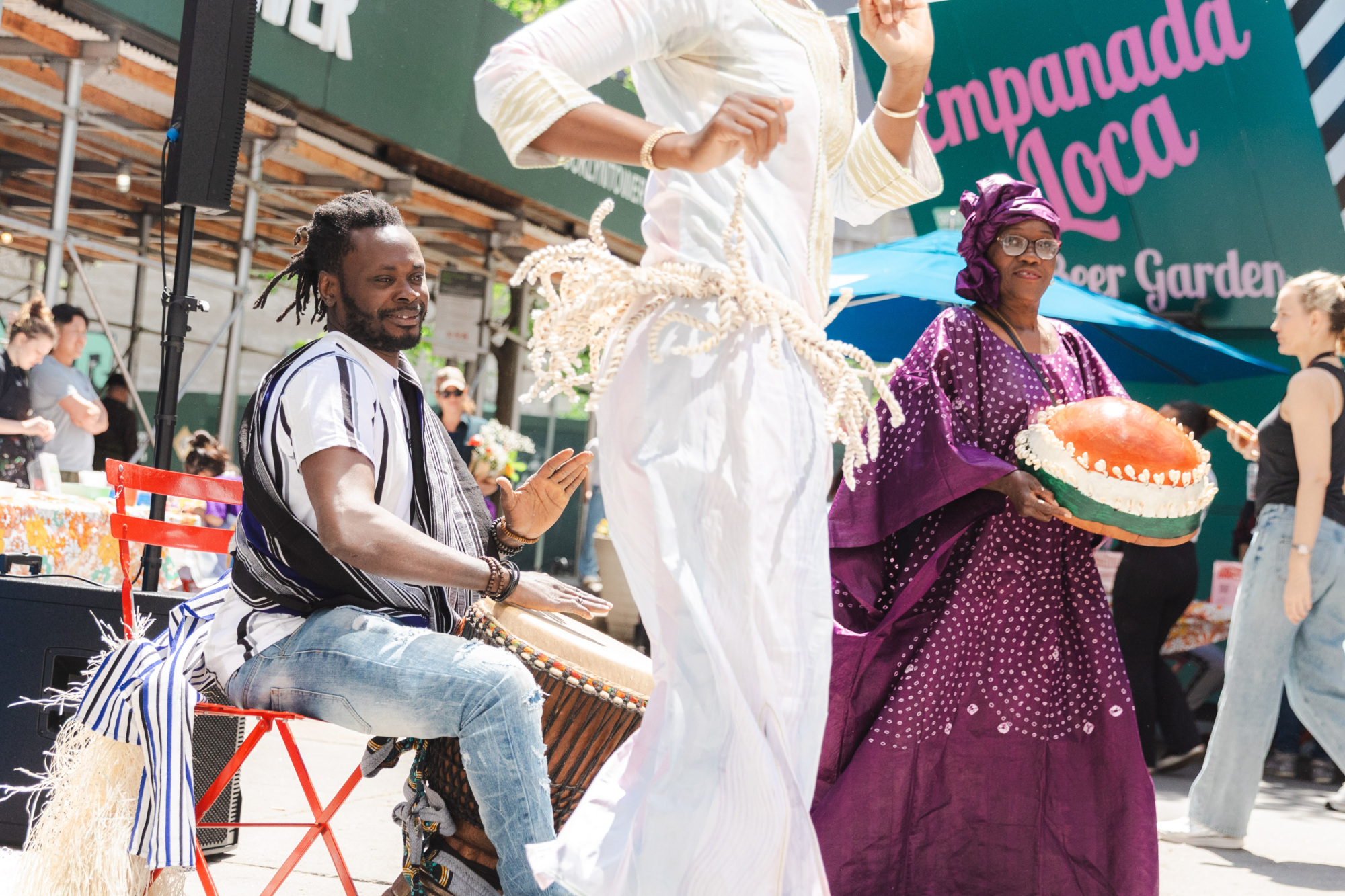 A man plays a drum while a person in white dances and a woman in a purple outfit holds a large decorative item at an outdoor event.
