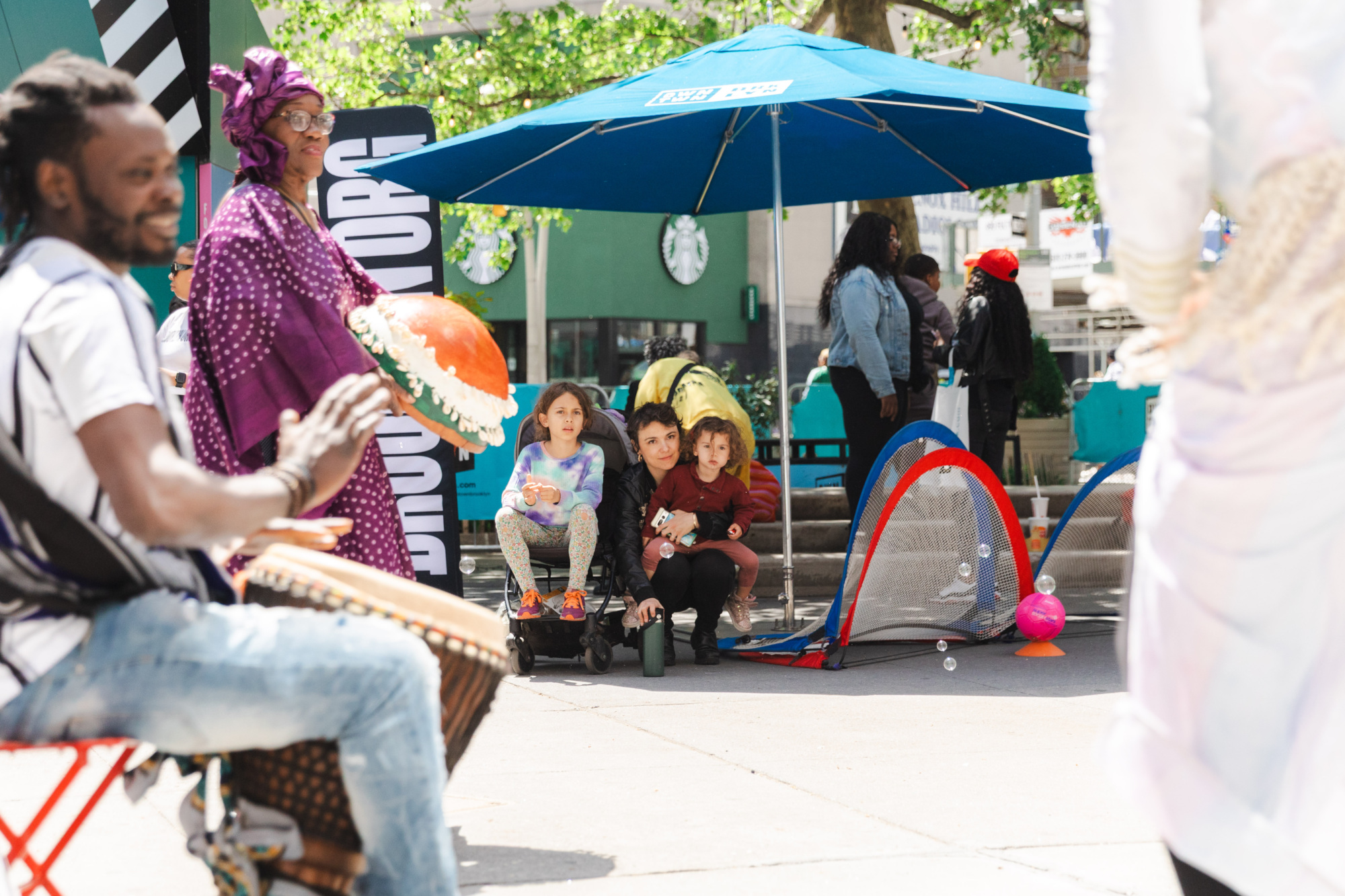 People gather outdoors for an event; a drummer plays in the foreground, children crouch near small soccer goals, and adults stand nearby under a blue umbrella.