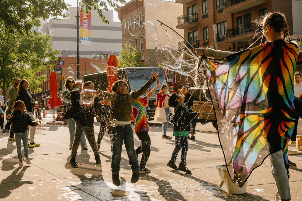 Children play and jump among bubbles on a city street while an adult in butterfly wings creates bubbles; people and buildings are visible in the background.