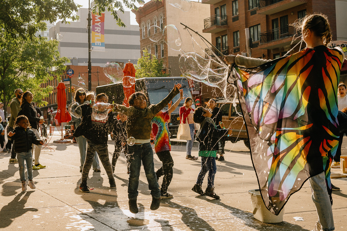 Children play and jump among bubbles on a city street while an adult in butterfly wings creates bubbles; people and buildings are visible in the background.