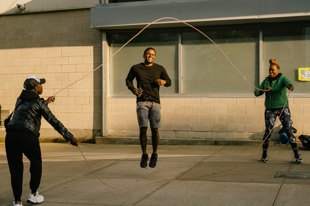 Three people are outside; two are turning a long jump rope while the third person is jumping in the middle, smiling.