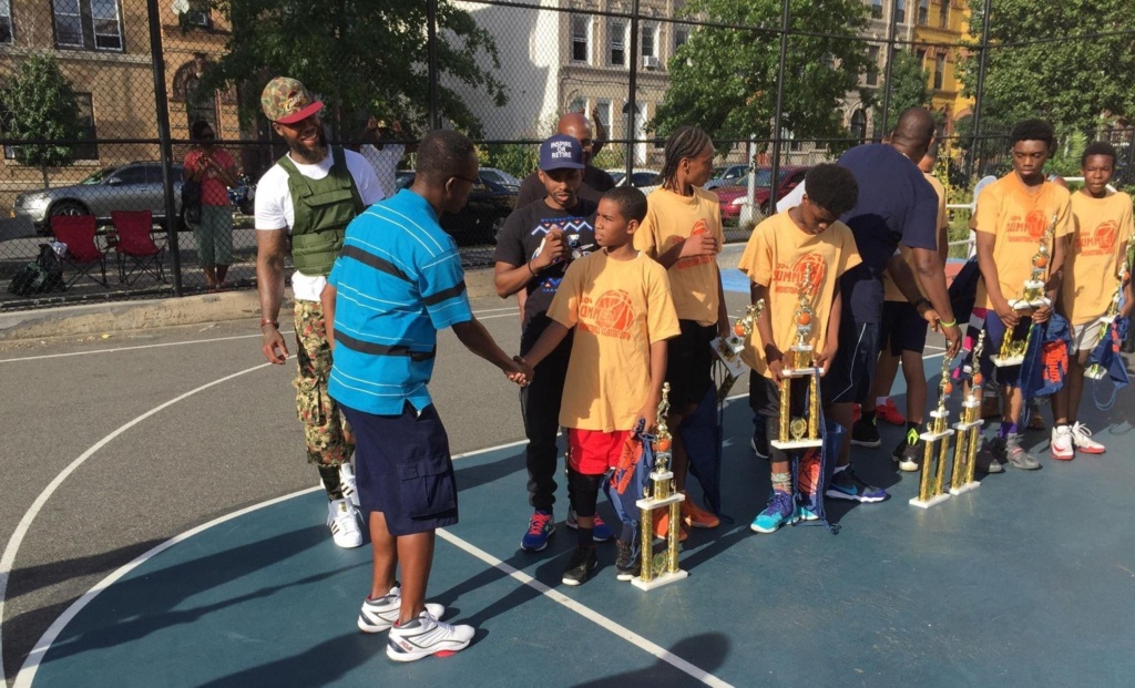 A group of boys on a basketball court hold trophies while a man in blue shakes hands with one boy; other adults stand nearby.