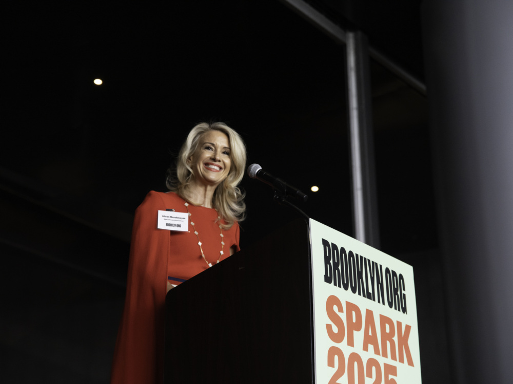 A woman stands at a podium with a microphone, wearing a red outfit and smiling, with a sign reading "BROOKLYN.ORG SPARK 2025" attached to the front.