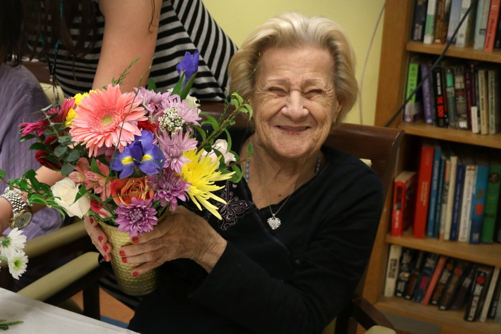 An elderly woman sitting and smiling while holding a colorful bouquet of flowers; another person’s arm helps arrange the bouquet. Bookshelves are visible in the background.