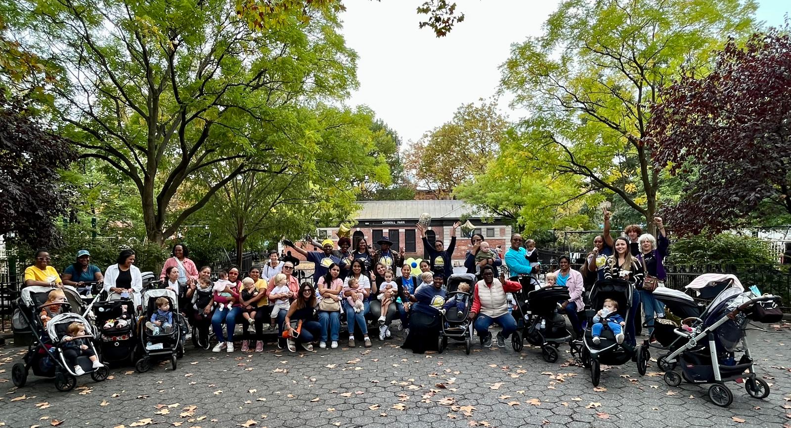 A large group of adults and children, many with strollers, gather outdoors in a park surrounded by trees and autumn foliage.